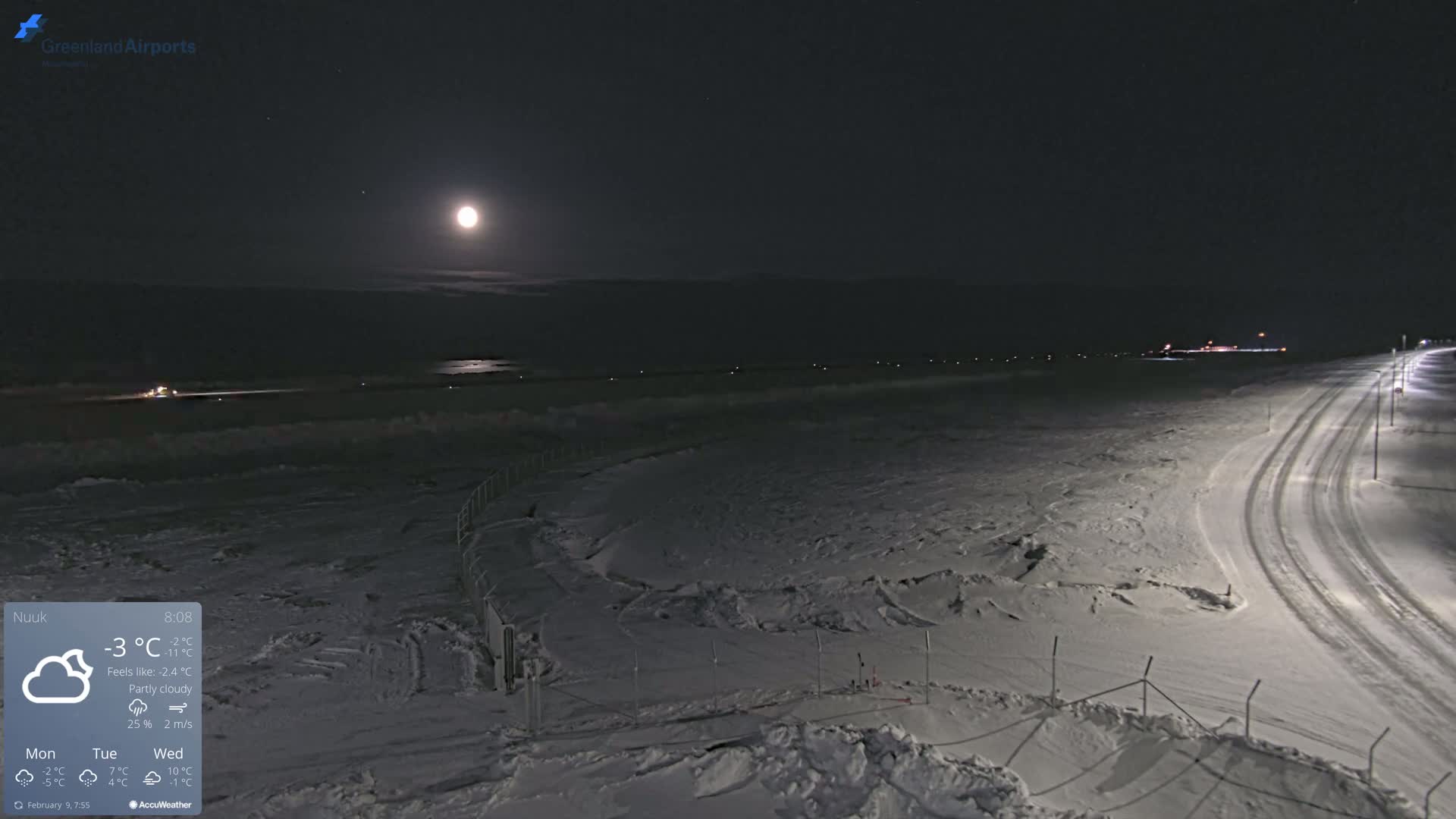A bright moon illuminates a snow-covered landscape at night, with a road and airport runway visible in the distance under partly cloudy conditions.