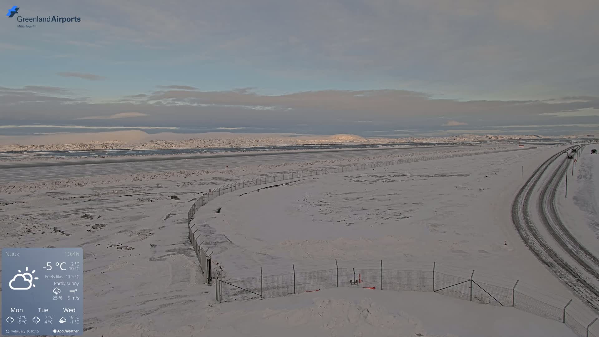 A snow-covered landscape with a road and fence, under partly sunny skies.