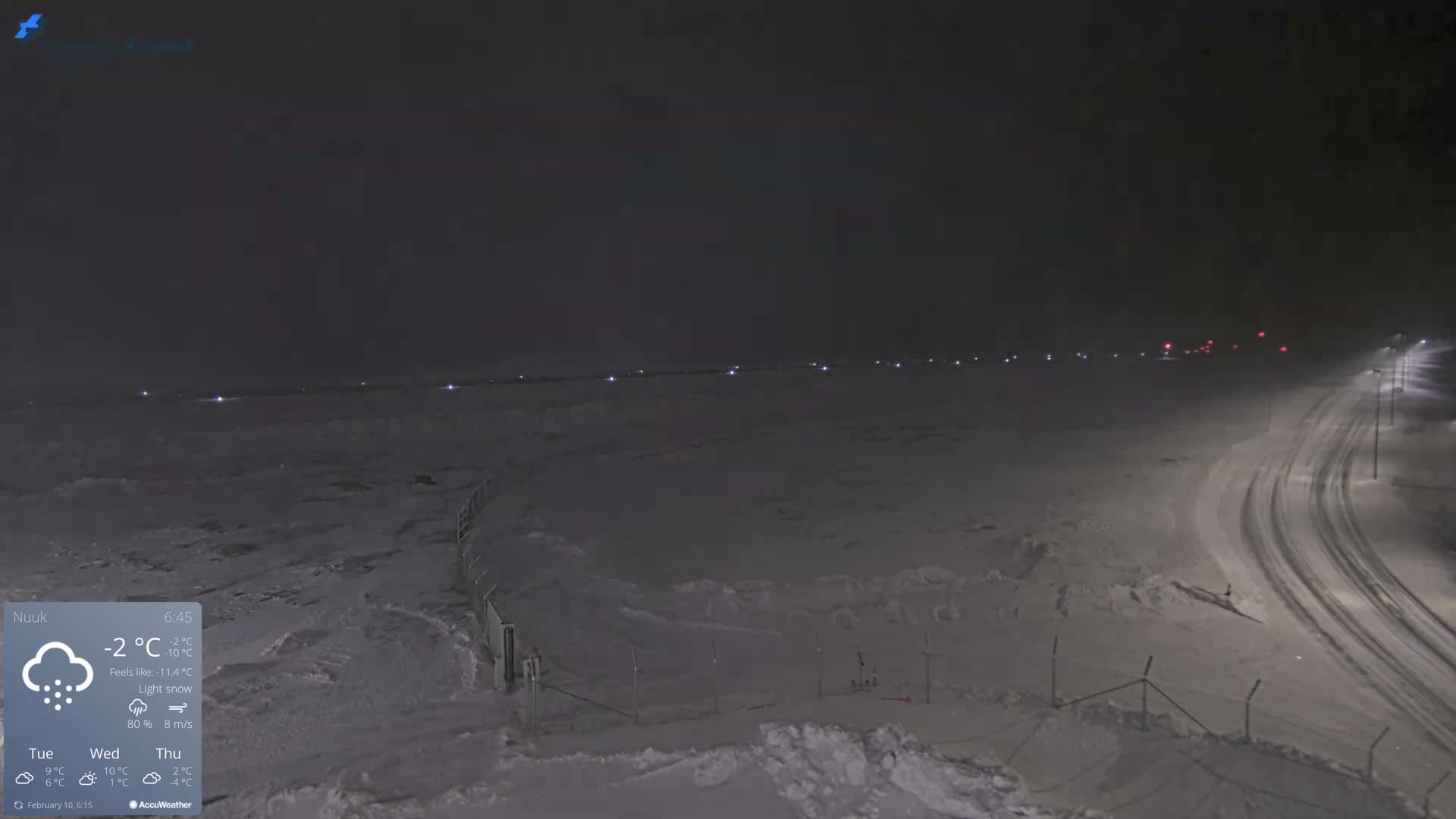 A snow-covered landscape at night is illuminated by distant lights and streetlights along a curving road, with light snow falling.