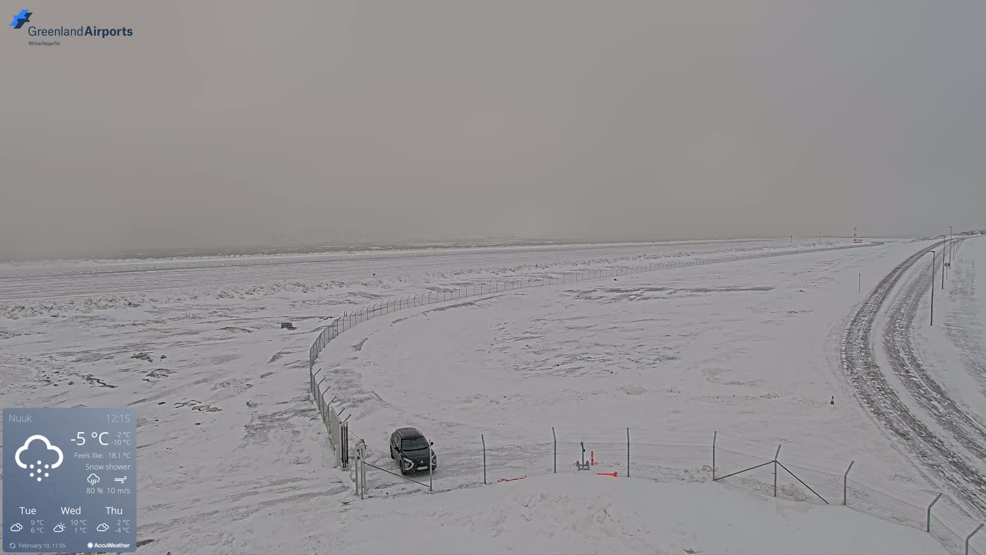 A snow-covered landscape with a car parked near a fence, overlooking a partially frozen sea under a gray, snowy sky.