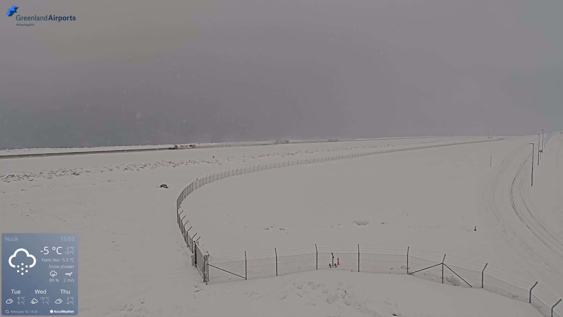 A snow-covered landscape with a chain-link fence and tire tracks is visible under a light snow shower.