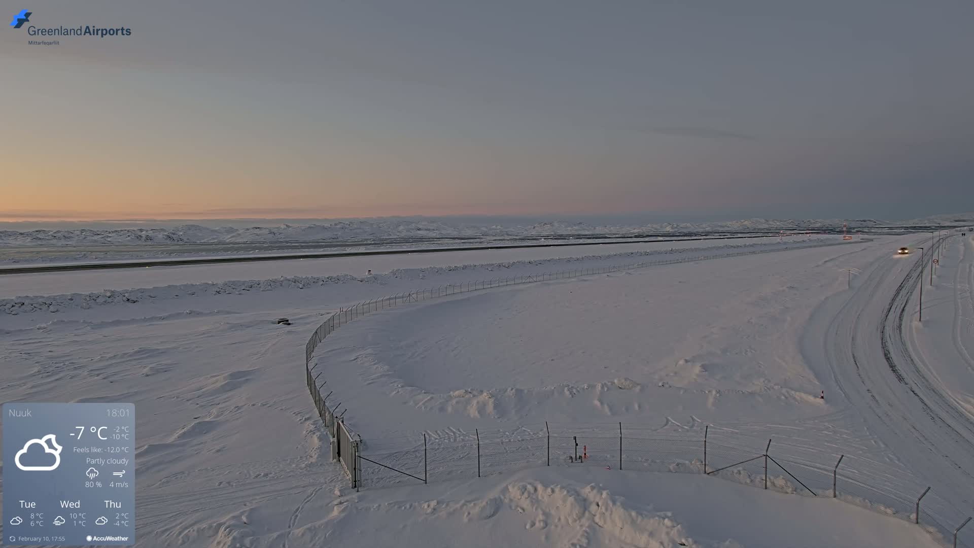 A snow-covered airport runway and surrounding area are visible under a partly cloudy sky during twilight.