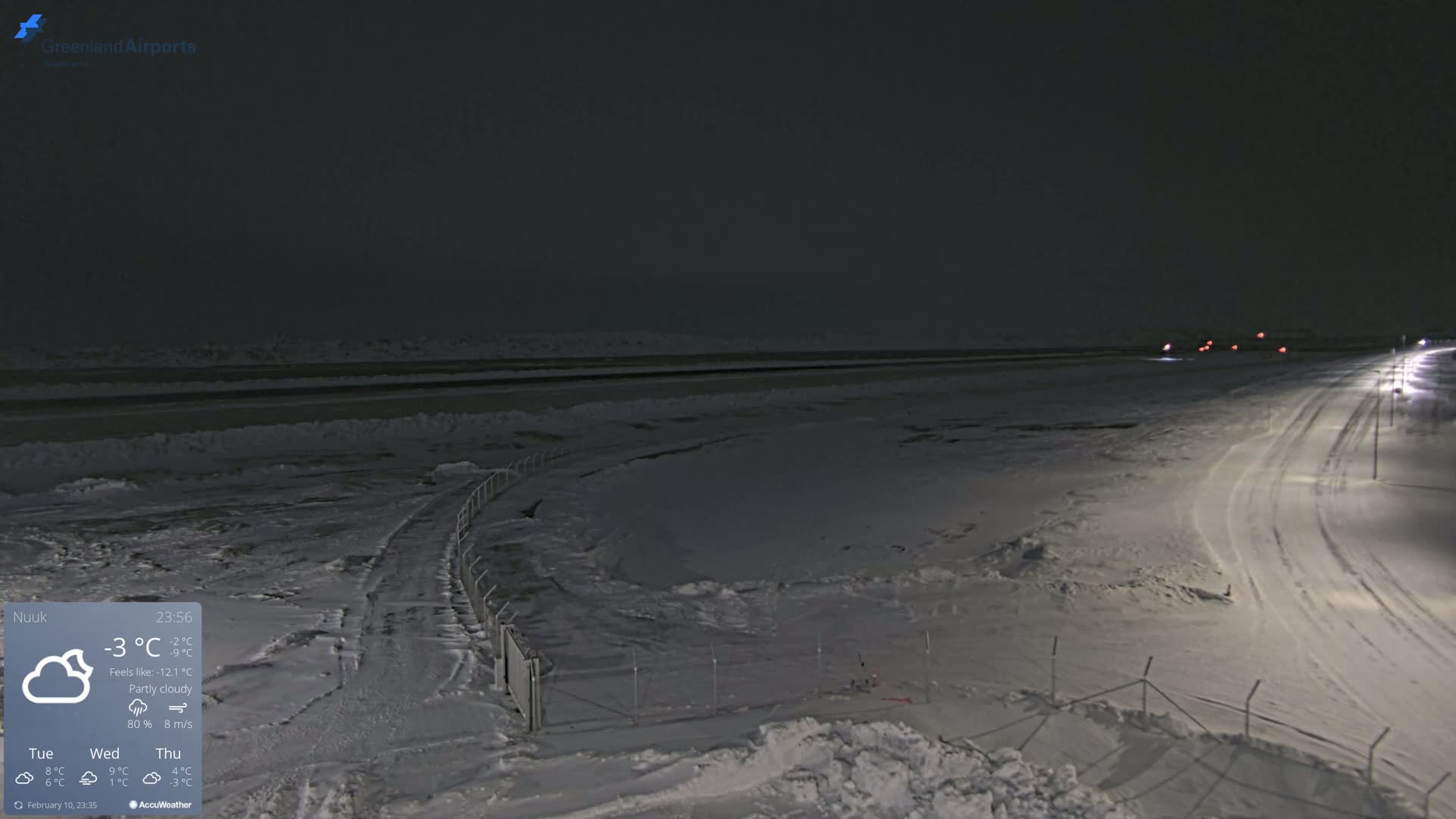 A snow-covered landscape at night, under partly cloudy skies, shows a road with tire tracks leading to a distant, dimly lit area.