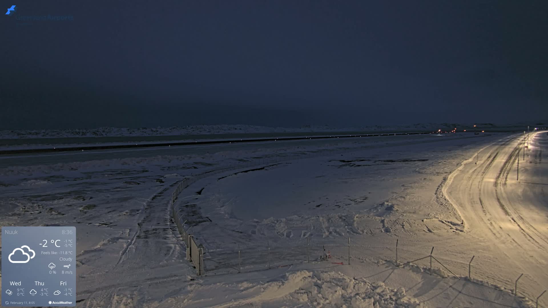 A snow-covered landscape at night, under cloudy skies, shows a road curving away from the viewer toward a body of dark water and distant lights.