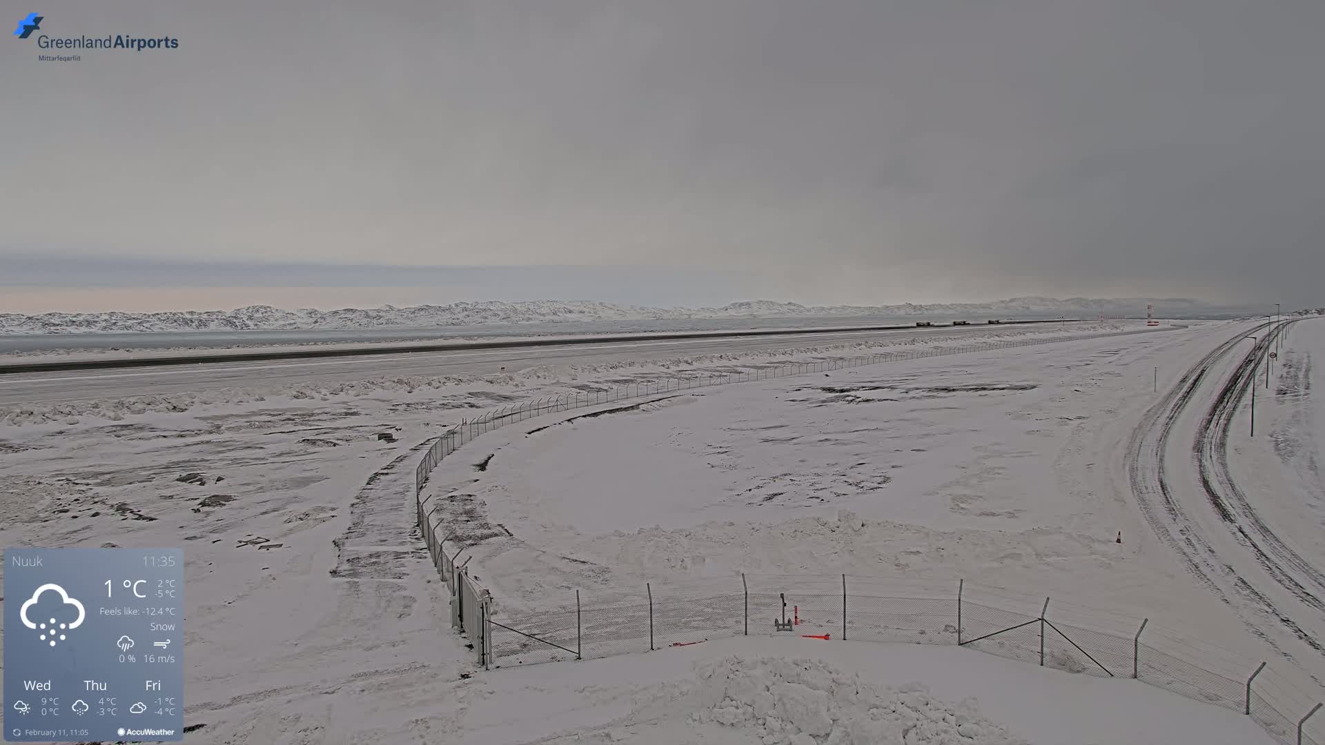 A snow-covered airport tarmac and runway extend to distant, snow-capped mountains under an overcast sky.