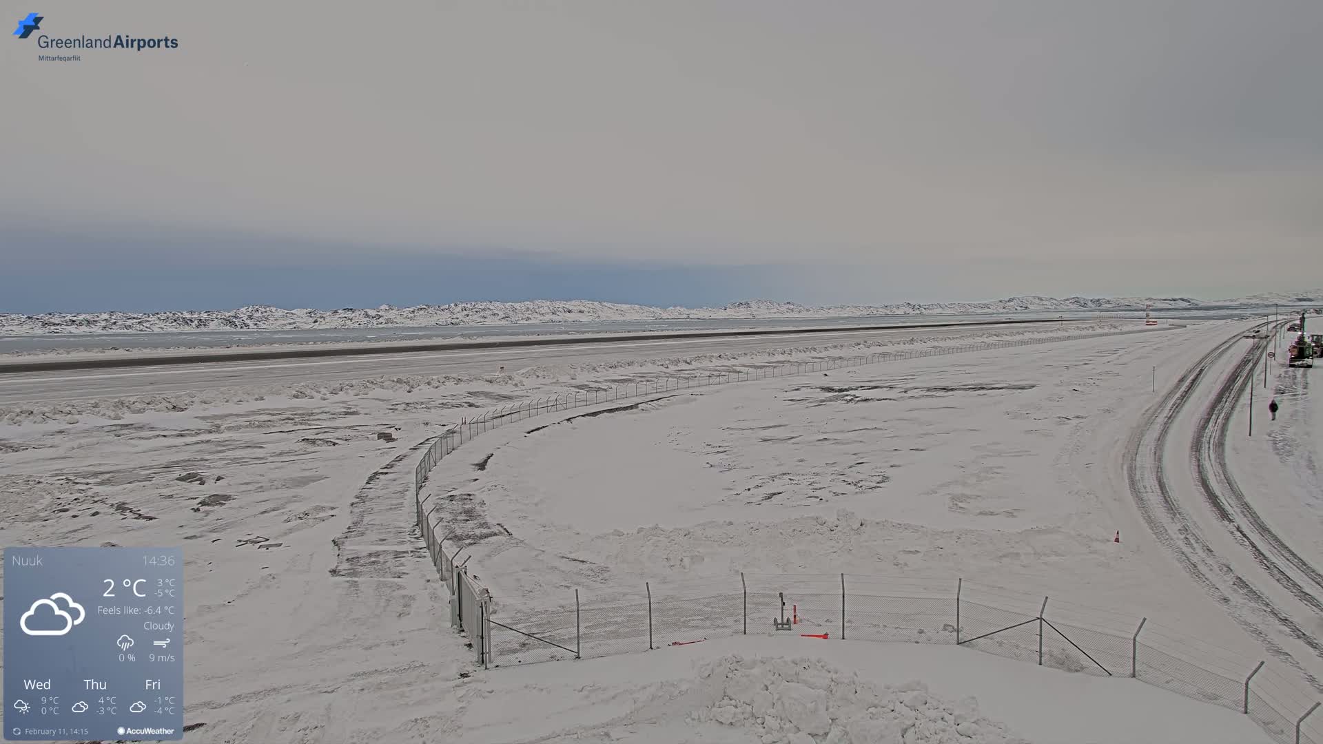A snow-covered airport runway and adjacent road stretch toward a distant, snow-capped mountain range under cloudy skies.