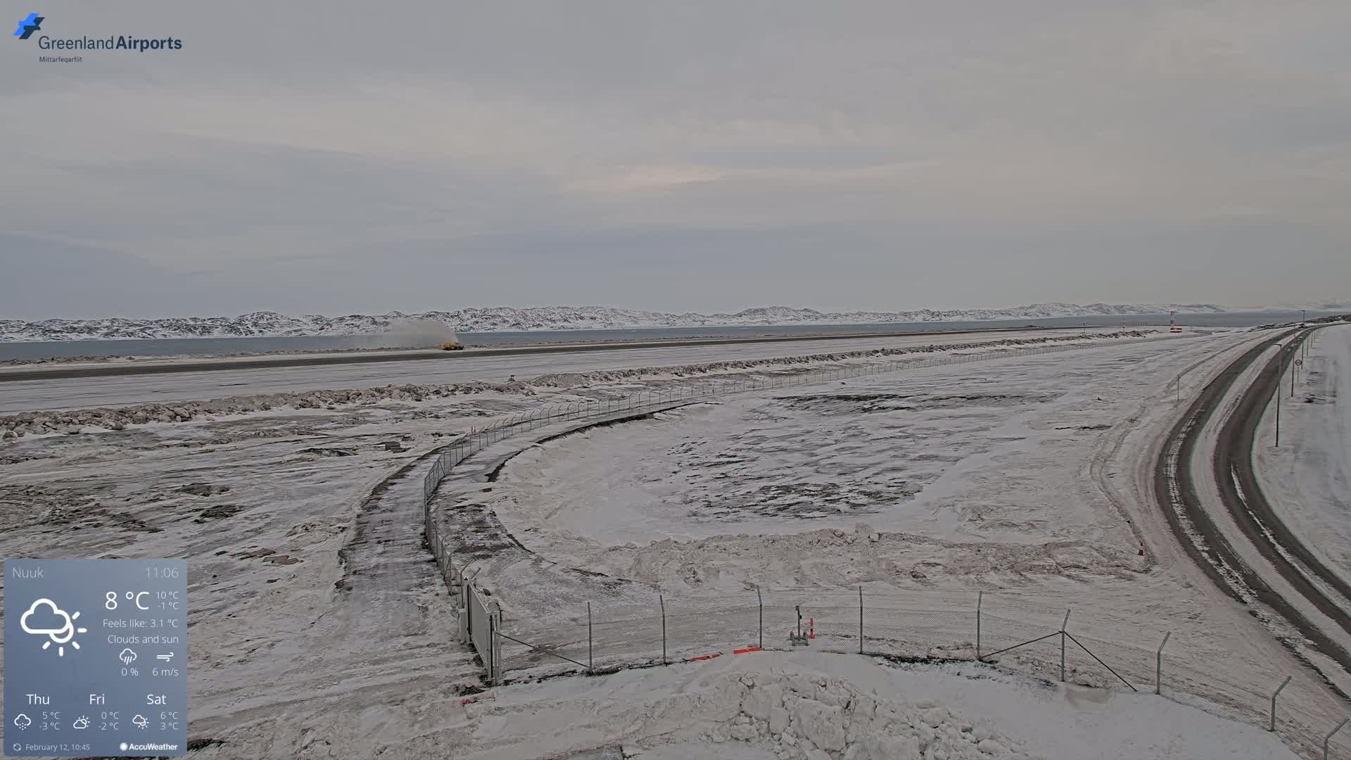 A snow-covered airport runway and adjacent roads are visible under cloudy skies, with distant snow-capped mountains in the background.