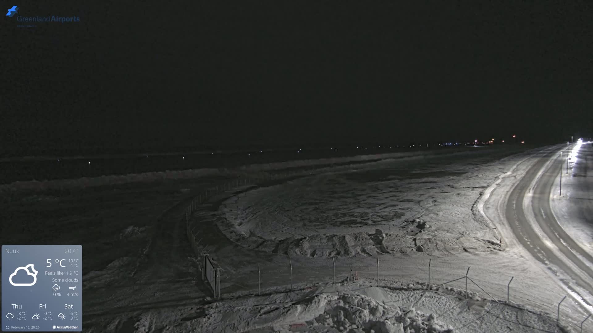 A snow-covered airfield at night under a dark sky with some clouds, illuminated by streetlights along a nearby road.