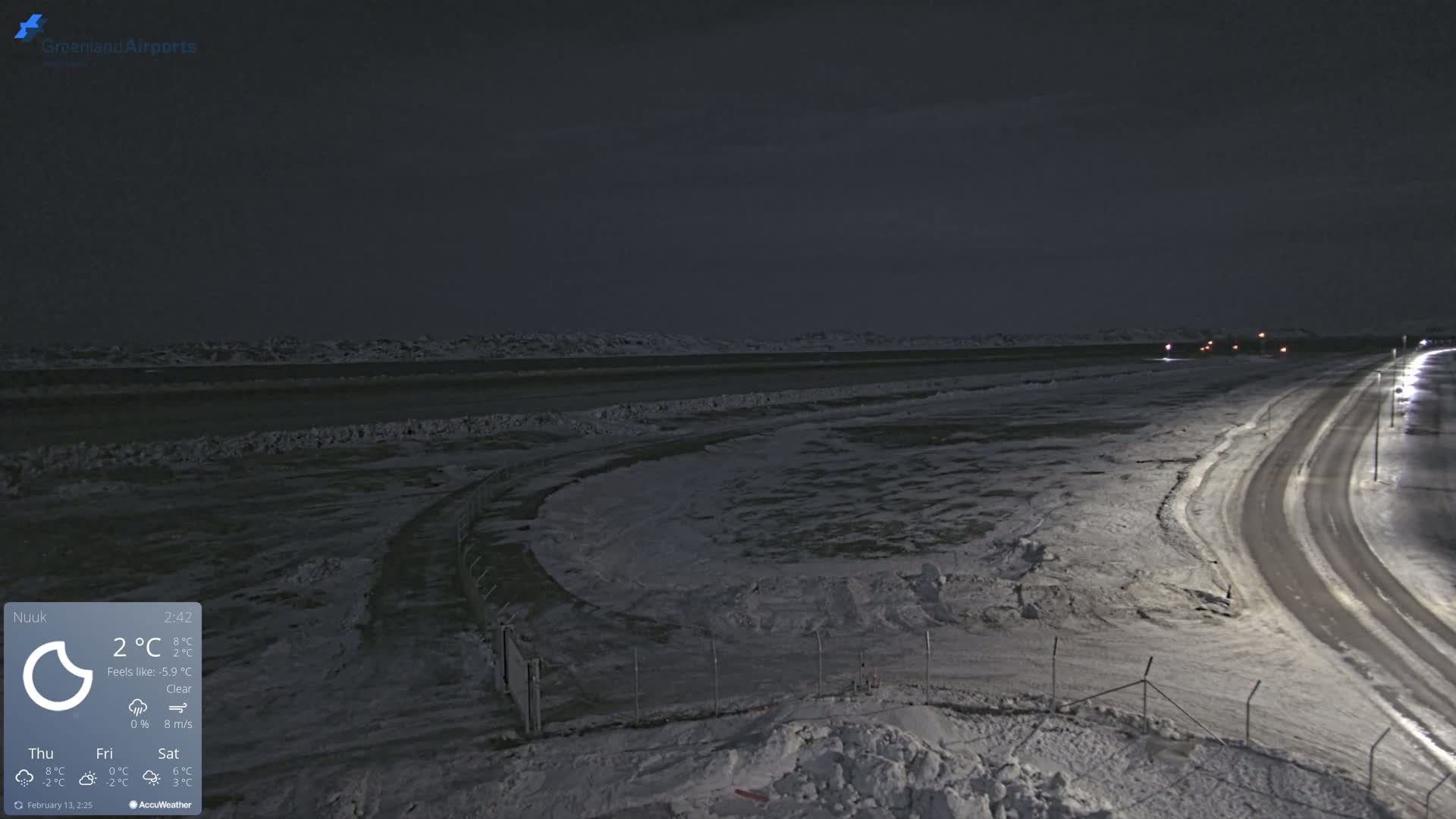 A snow-covered landscape at night, with a road curving toward the viewer, and a body of dark water in the distance under a clear sky.