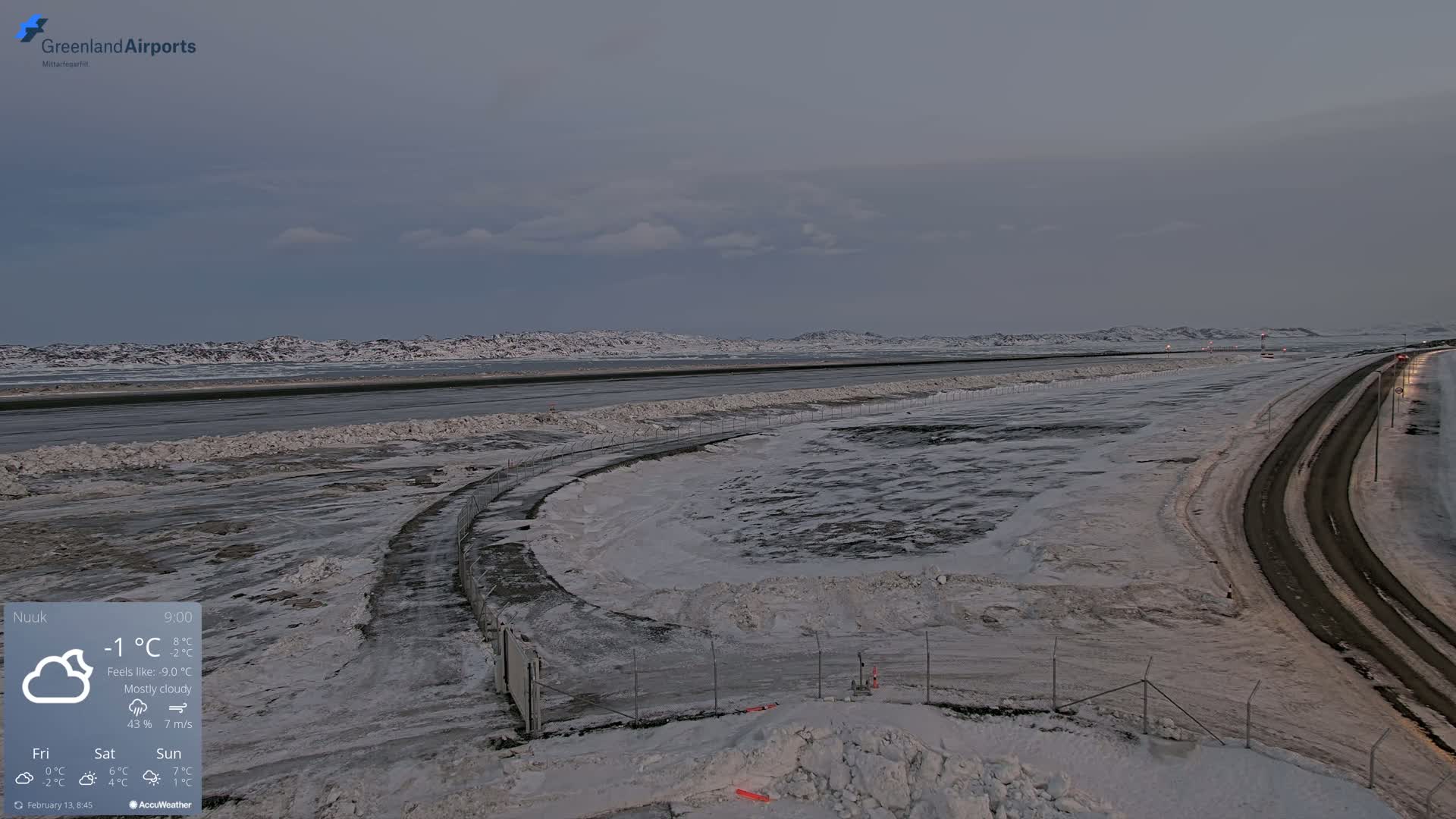 A mostly cloudy, snowy, and cold landscape shows a snow-covered airport runway and road, with distant snow-covered hills under a pale sky.