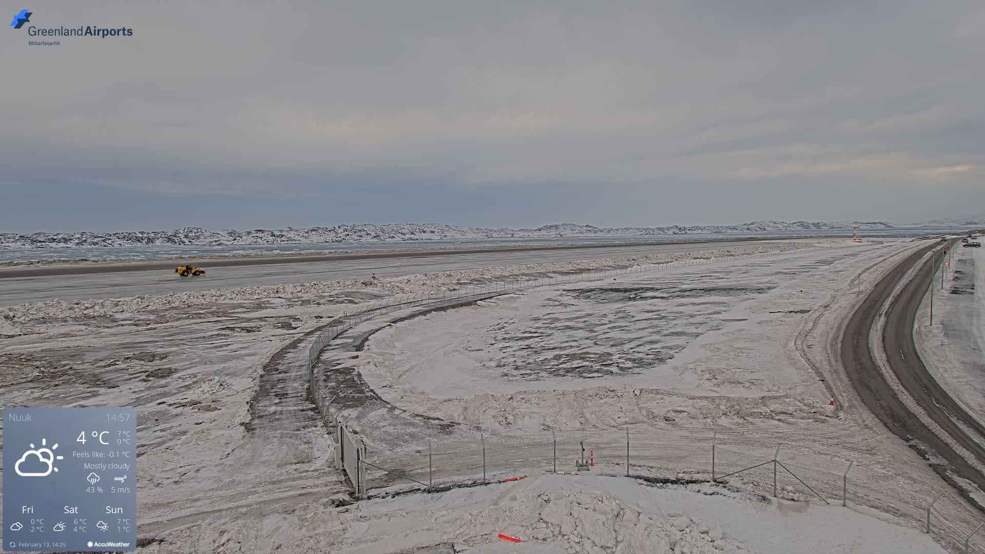 A mostly cloudy day shows a snow-covered airport with a road and a piece of heavy machinery visible in the distance.