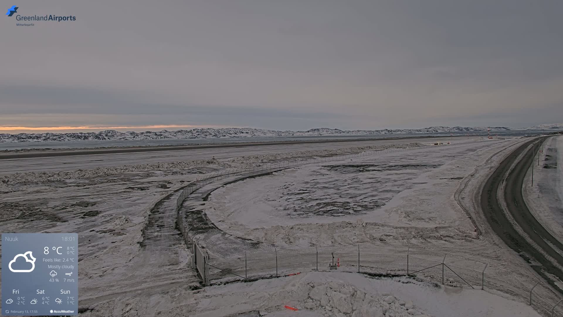 A mostly cloudy, snowy airfield with a road running alongside it, overlooks a snow-covered landscape and distant, snow-capped mountains under a pale sky.