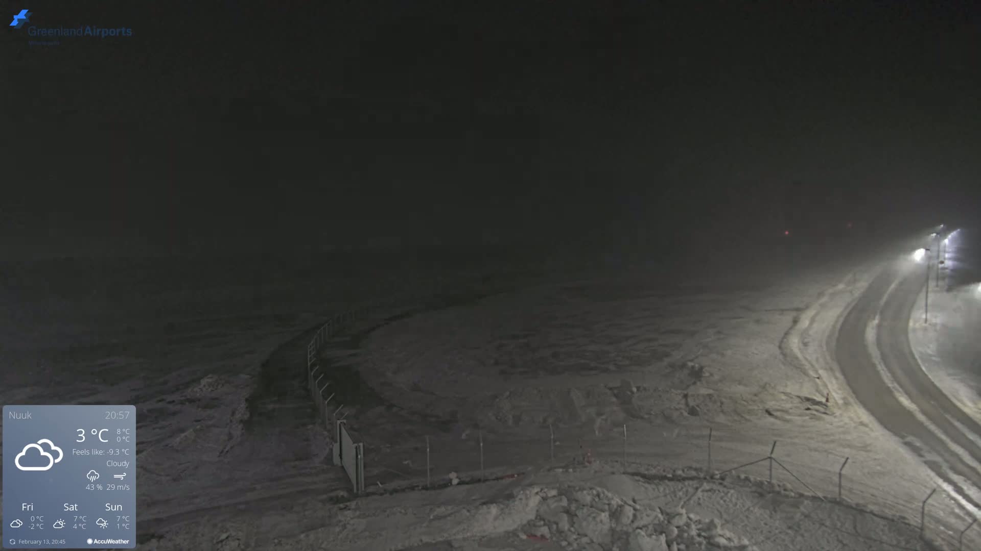 A snow-covered, dimly lit area with a road curving away from the viewer under a cloudy, dark night sky.