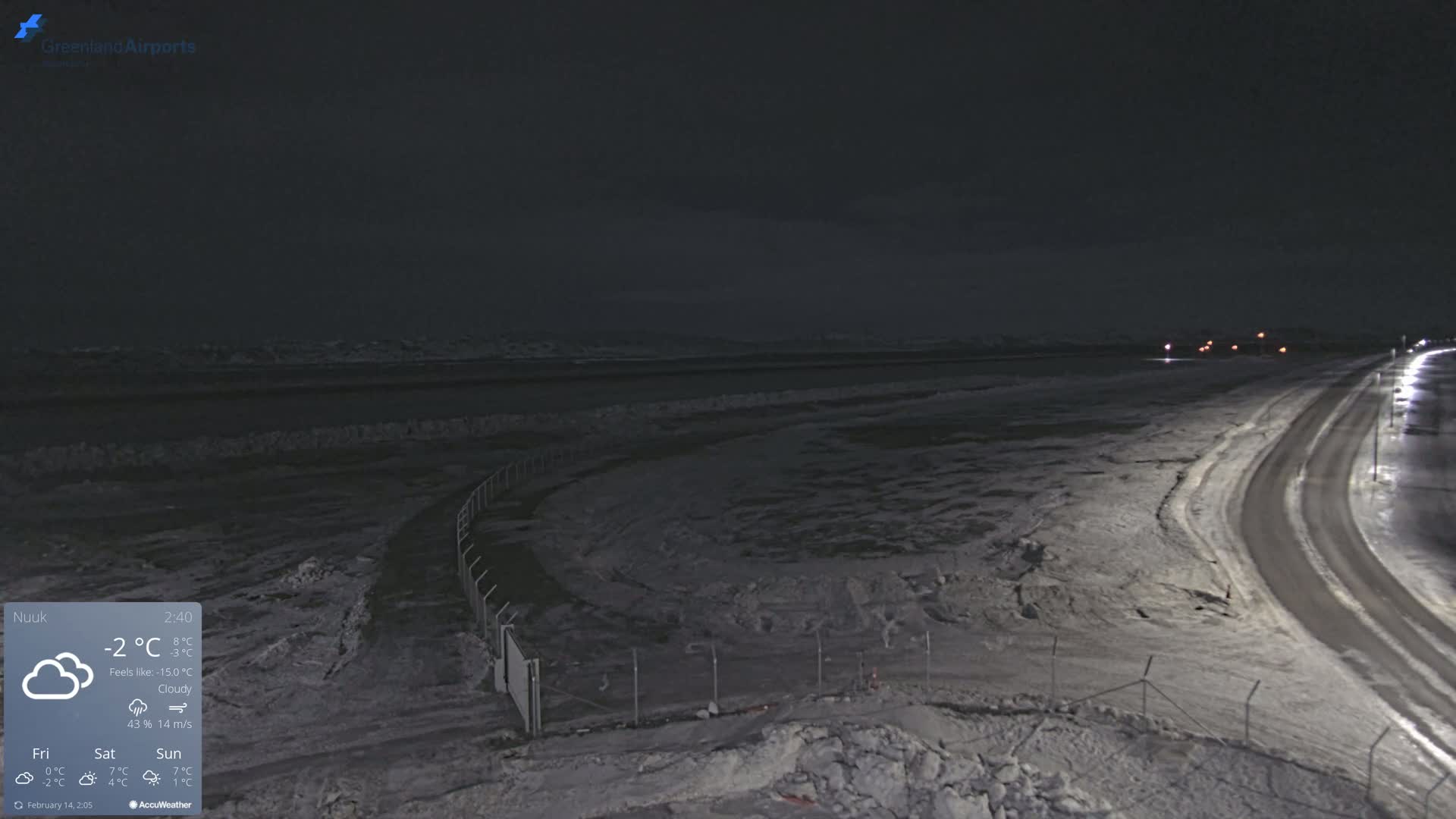 A snow-covered, dark landscape at night shows a road curving toward a body of water under a cloudy sky.