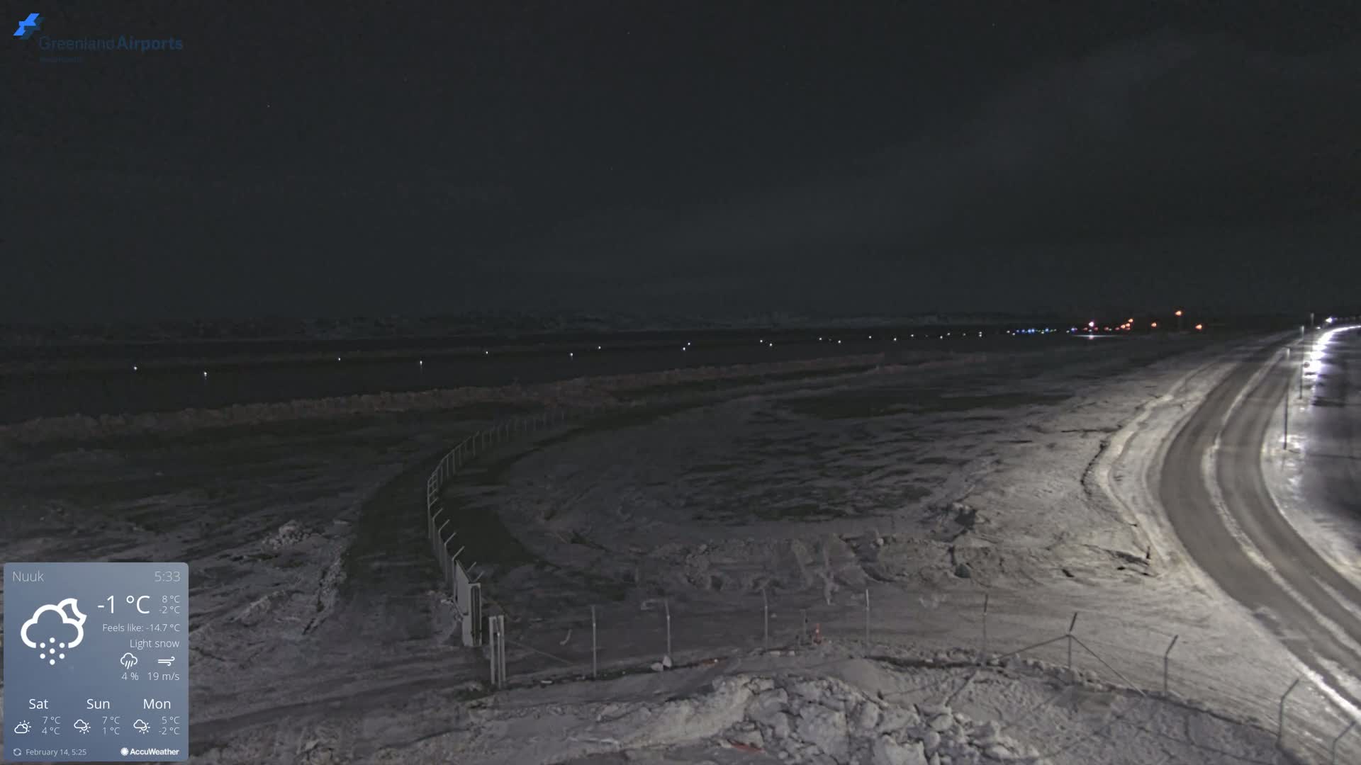 A snow-covered, dimly lit airfield at night with a road curving towards the background under light snowfall.