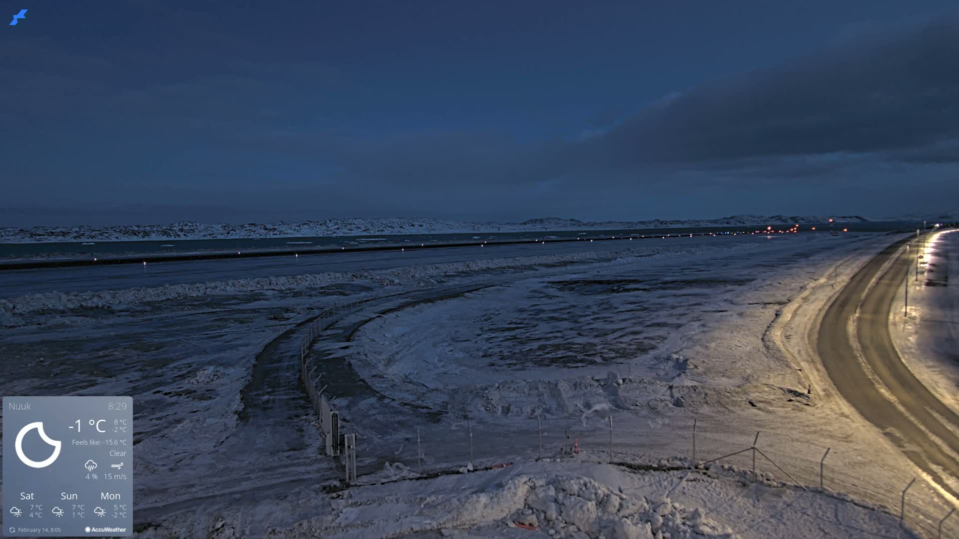 A snow-covered landscape at twilight shows a road curving toward a body of water with a distant, snow-covered mountain range under a clear, dark sky.