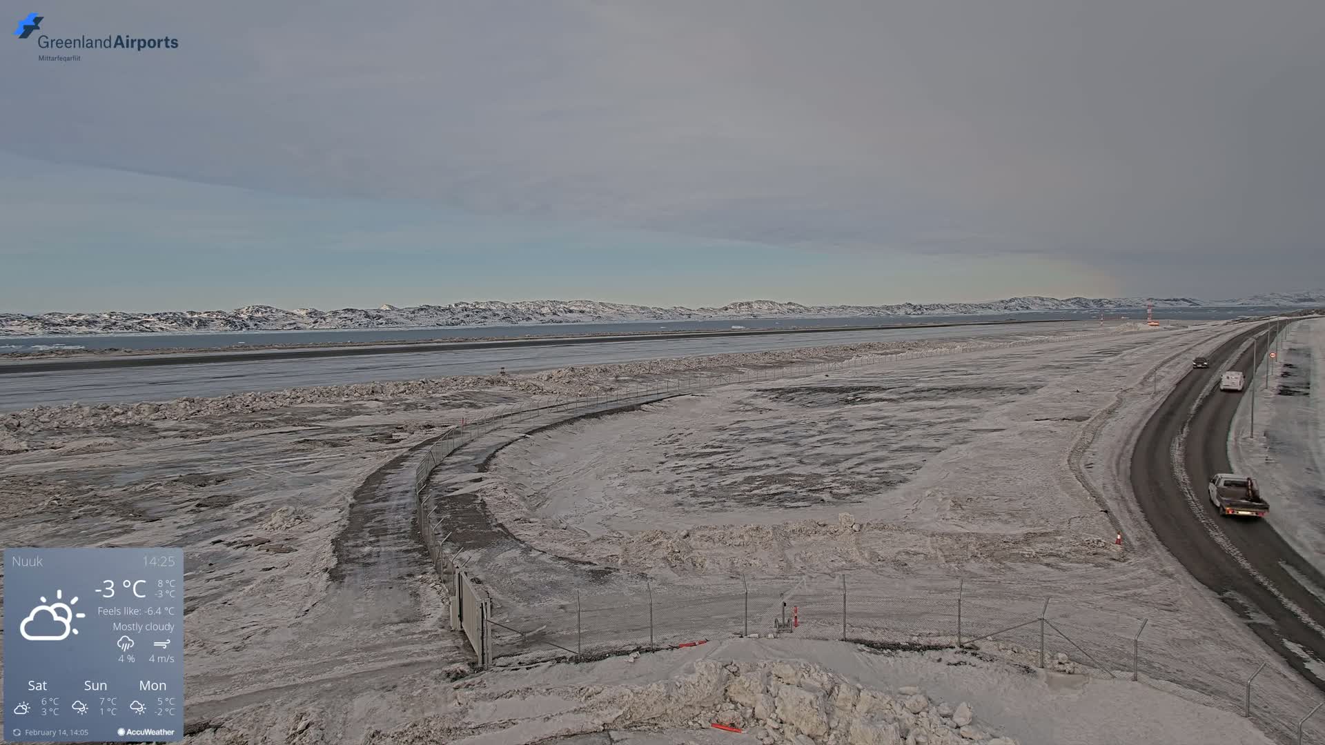 A mostly cloudy, cold day reveals a snow-covered airport with a road and a few vehicles visible, overlooking a frozen body of water and distant snow-capped mountains.