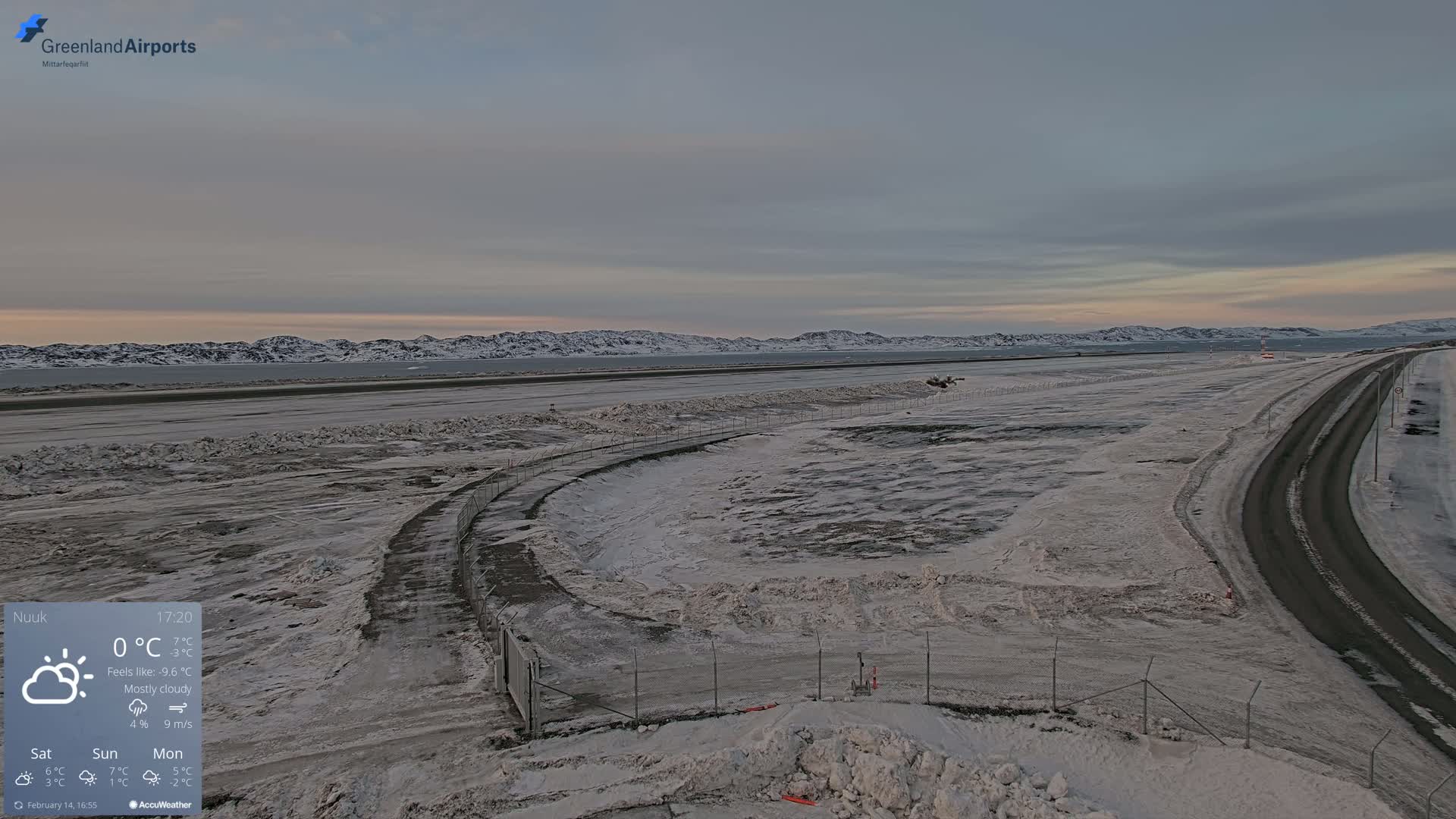 A mostly cloudy, snowy airfield with a road beside it and mountains in the distance.