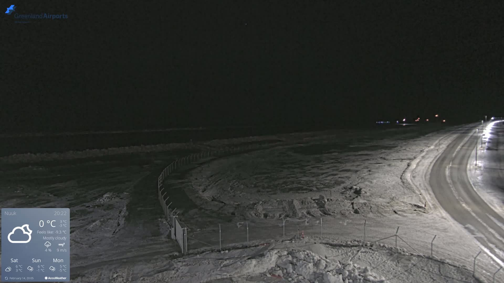A mostly cloudy night scene shows a snow-covered road curving alongside a fenced-in area, with a dark body of water visible in the background under a dark sky.