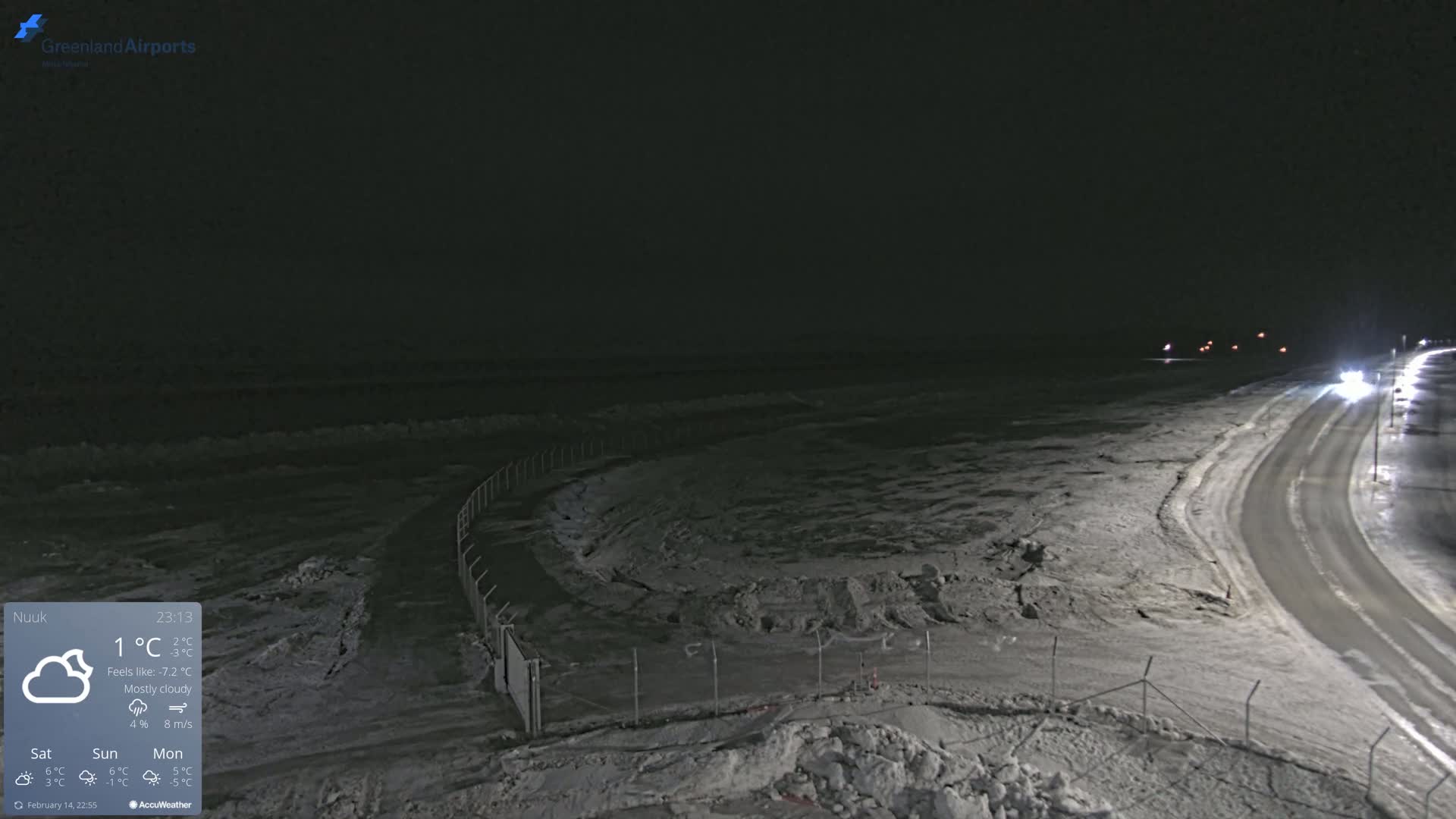 A nighttime, mostly cloudy scene shows a snow-covered landscape with a road curving towards a dark body of water under dim lighting.