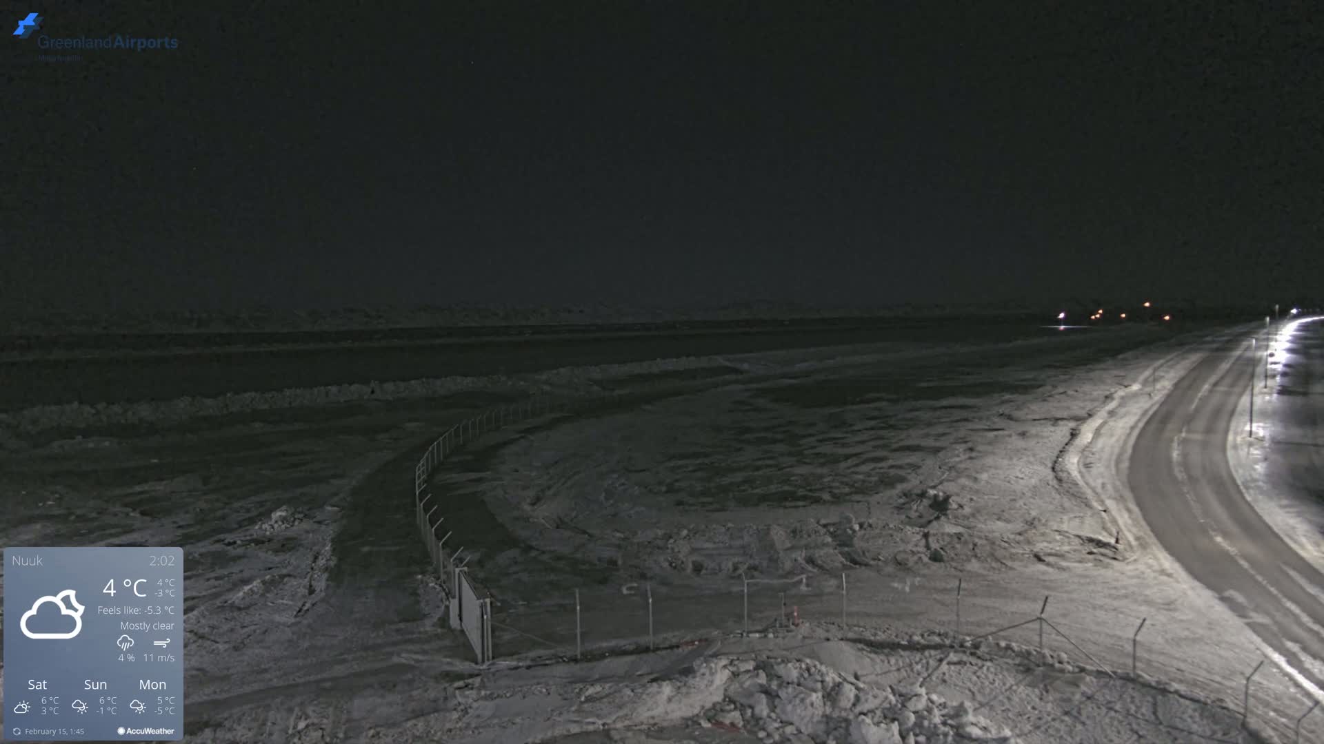 A mostly clear night scene shows a snow-covered road curving toward a body of water under a dark sky.