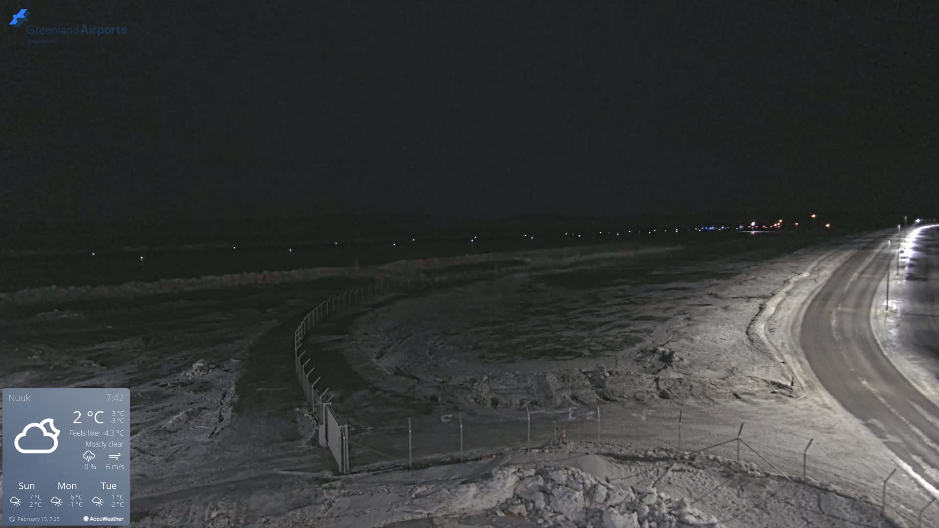 A snow-covered airfield is seen at night under mostly clear skies, with a road curving toward the right.