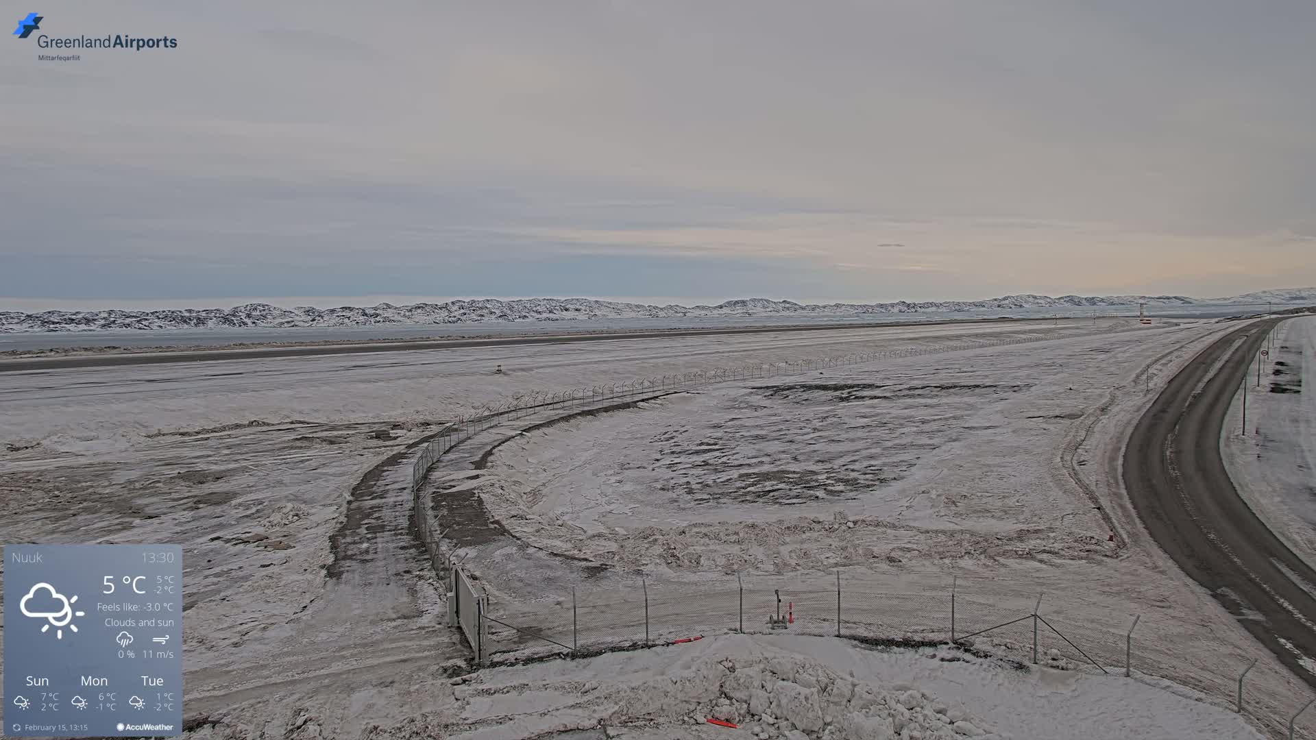 A snow-covered airport runway and road extend toward a distant, snow-capped mountain range under a cloudy sky.