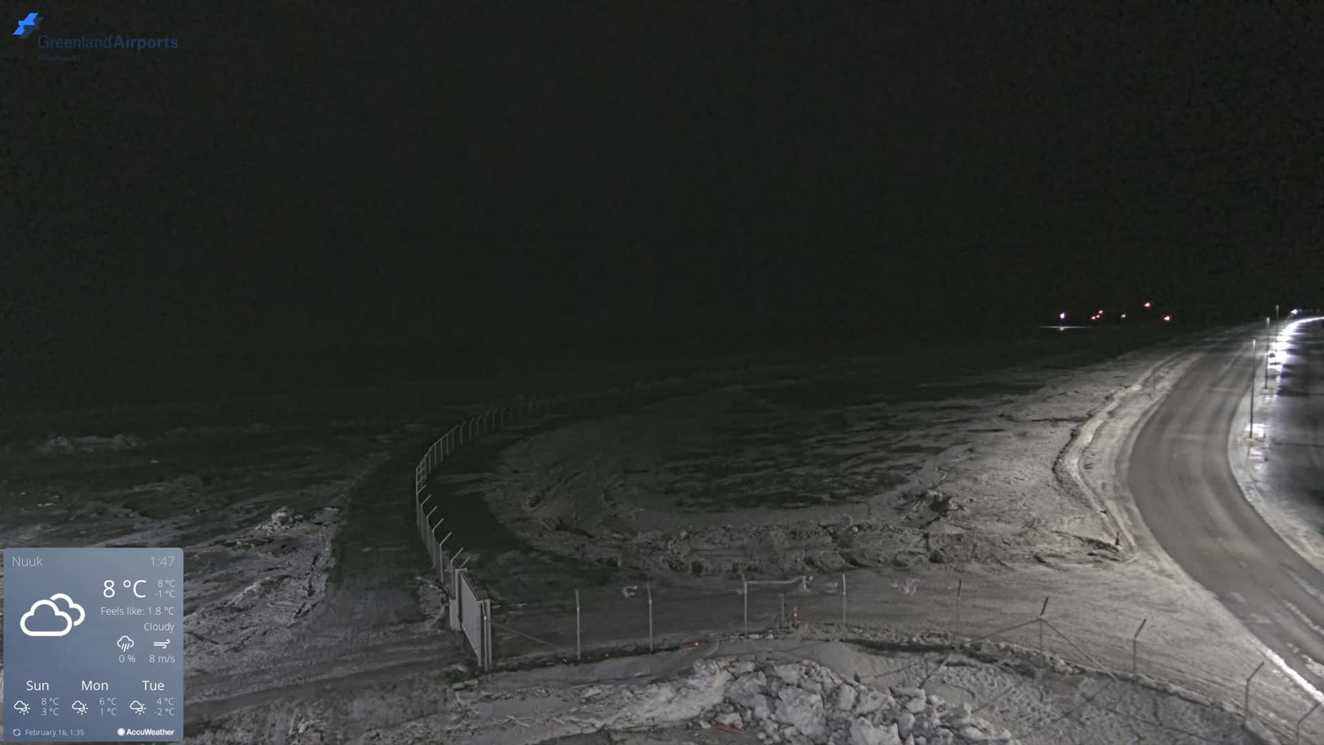 A snow-covered, dark nighttime scene shows a road curving away from the camera, bordered by a fence and illuminated by streetlights.