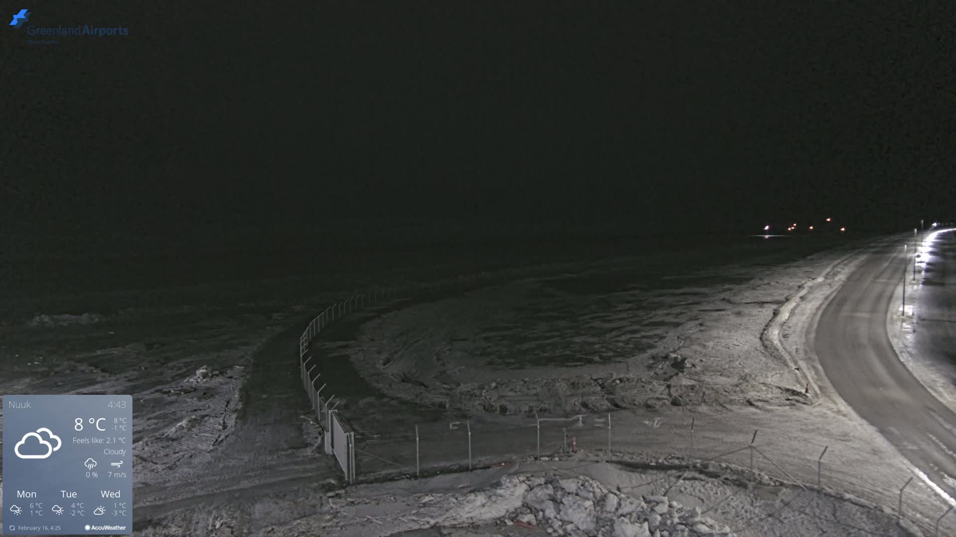 A nighttime, cloudy scene of a snow-covered road curving alongside a fenced area, with faint lights visible in the distance.