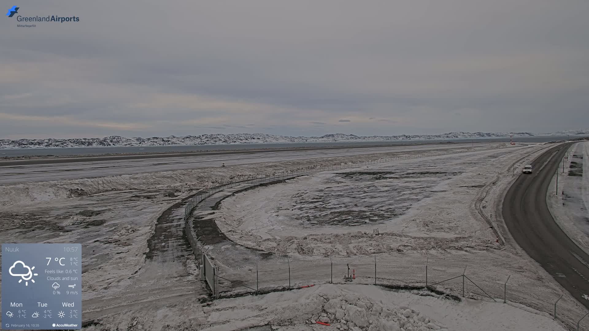 A snow-covered airport runway and road are visible under a cloudy sky, with snow-capped mountains in the distance.