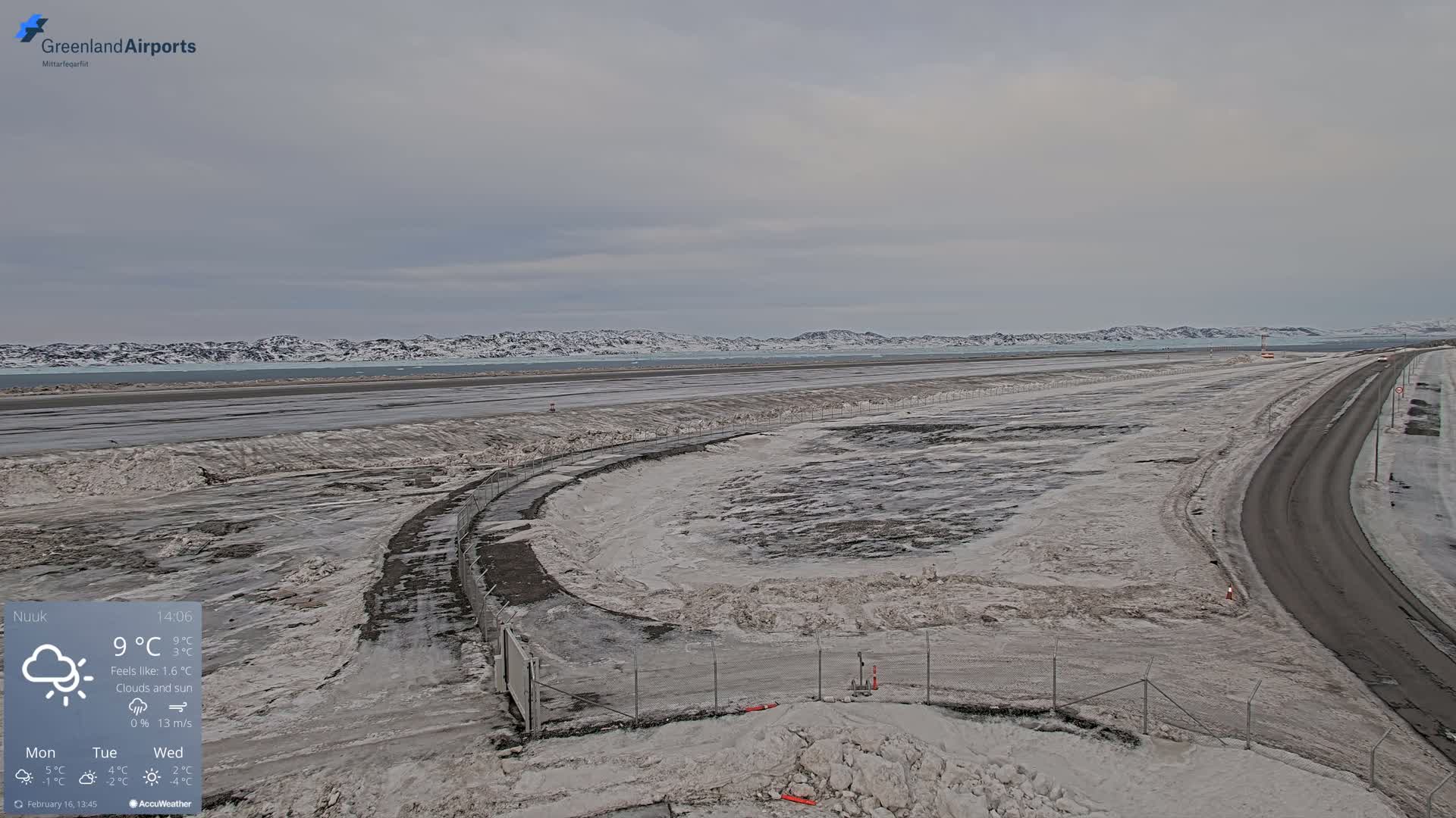 A snow-covered airport tarmac and access road extend towards a distant, snow-capped mountain range under a cloudy sky.