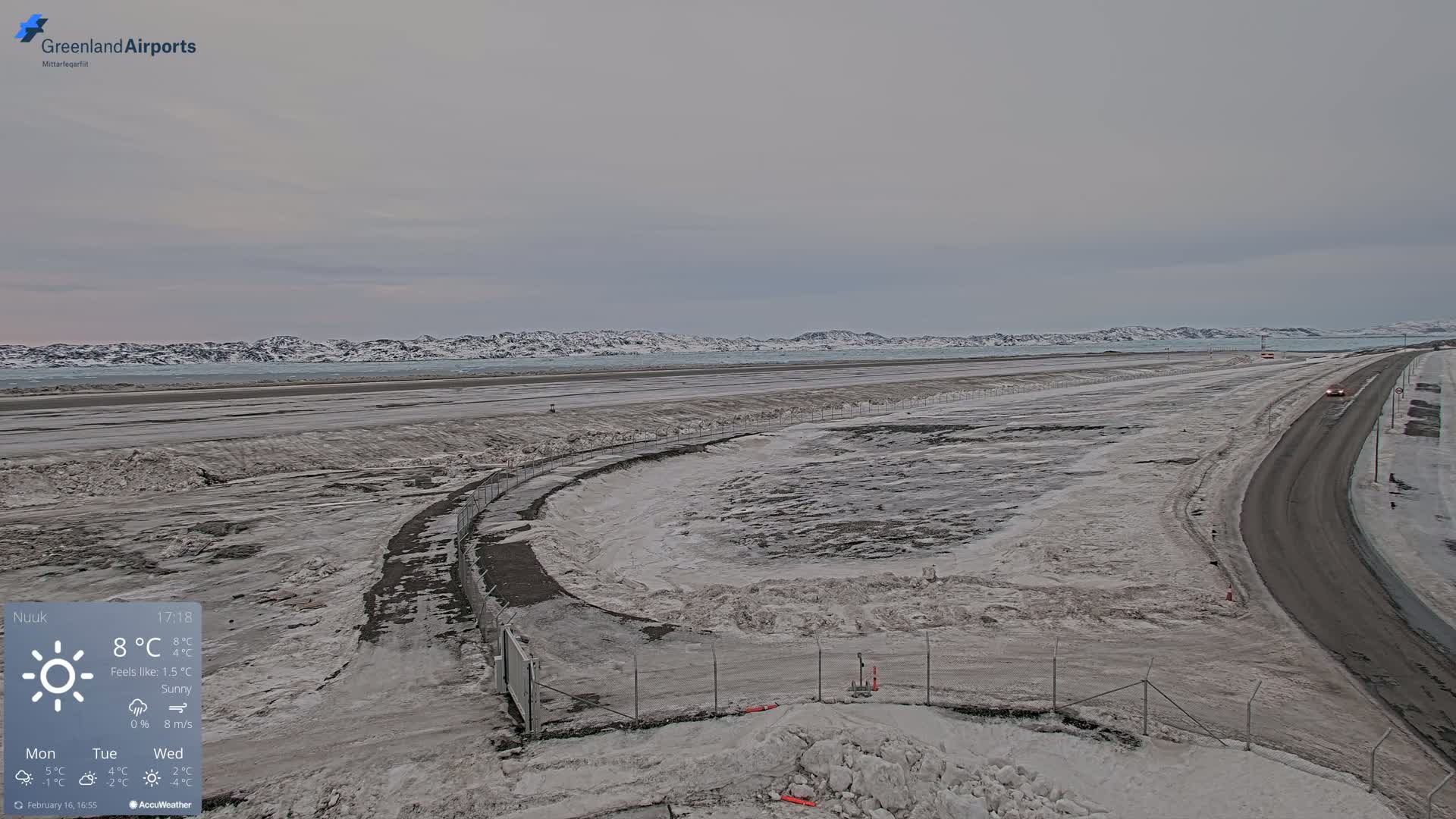 A snowy, sunny landscape featuring a road and airport runway next to a body of water with snow-covered mountains in the background.