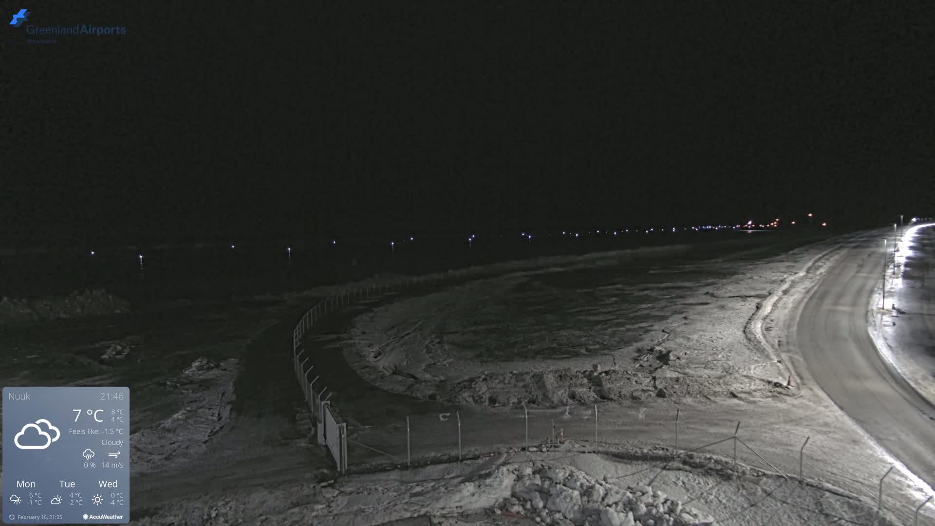 A snow-covered, nighttime airport scene with a road curving towards the right, under cloudy skies.
