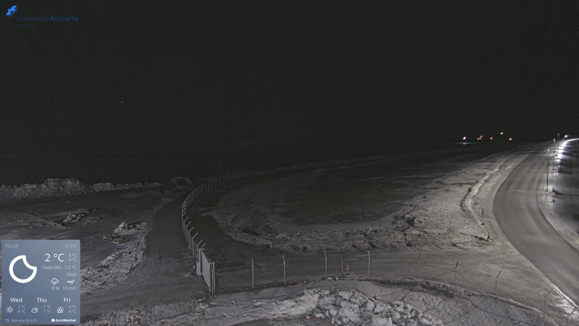 A snow-covered, dark, nighttime scene shows a road curving towards some distant lights, with a chain-link fence alongside a snow-dusted expanse of land.