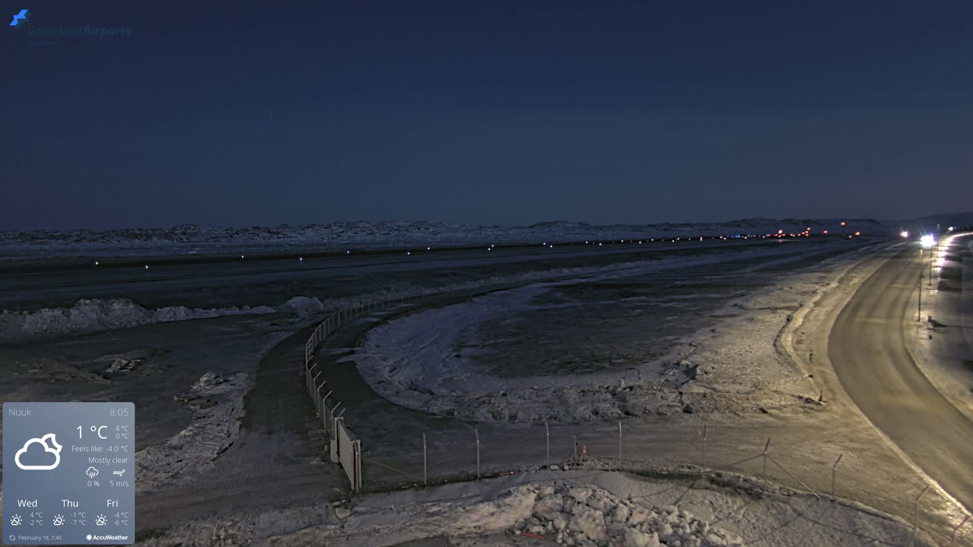 A mostly clear night scene shows a snow-covered airfield with a road curving away to the right under a dark blue sky.