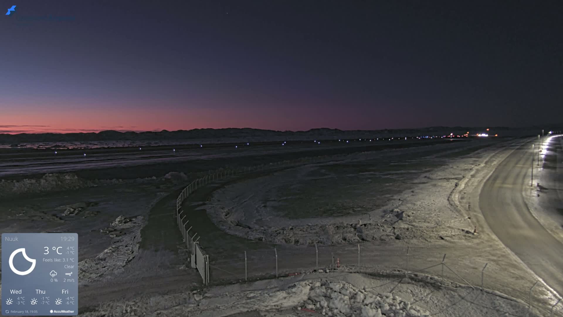 A snow-covered landscape at twilight, with a road curving toward a distant, dimly lit area and a faint pink hue on the horizon.