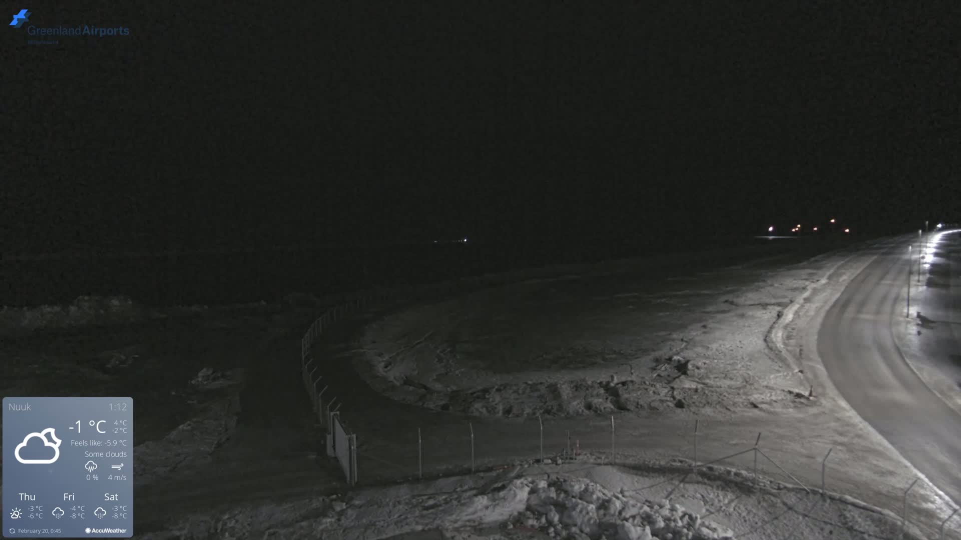 A nighttime, snow-covered scene shows a road curving toward a body of water under a partly cloudy sky.