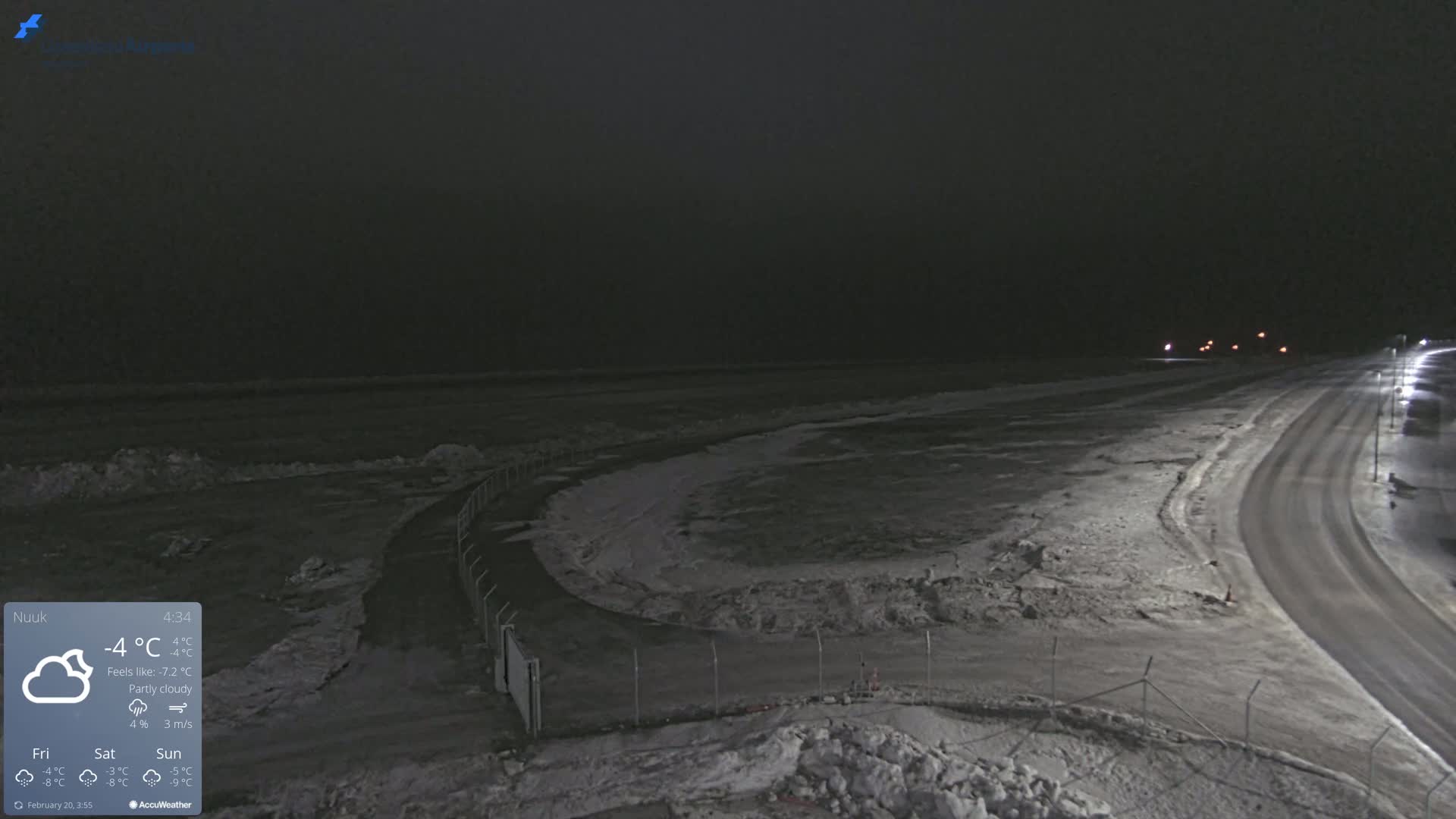 A nighttime, partly cloudy scene shows a snow-covered road curving past a fenced-in area next to a dark body of water with ice formations.