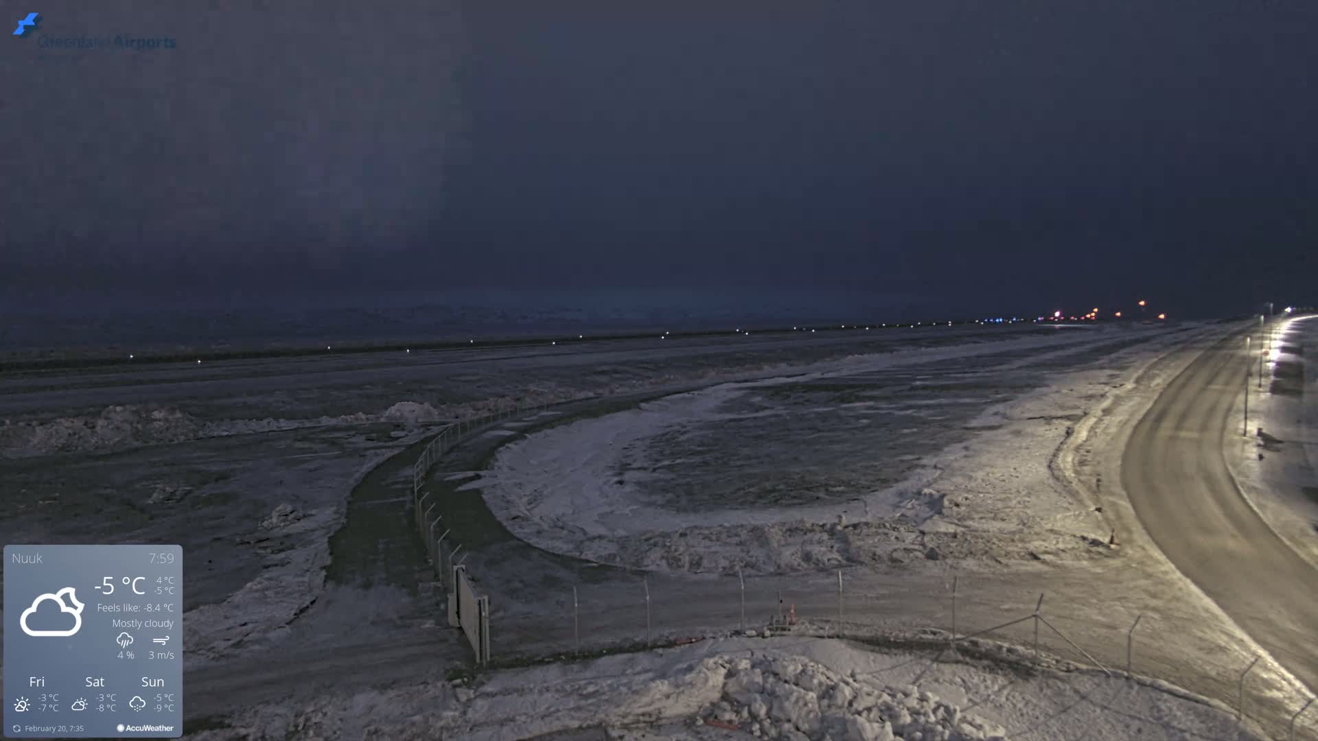A mostly cloudy night scene shows a snow-covered airport runway and road under dim lighting.