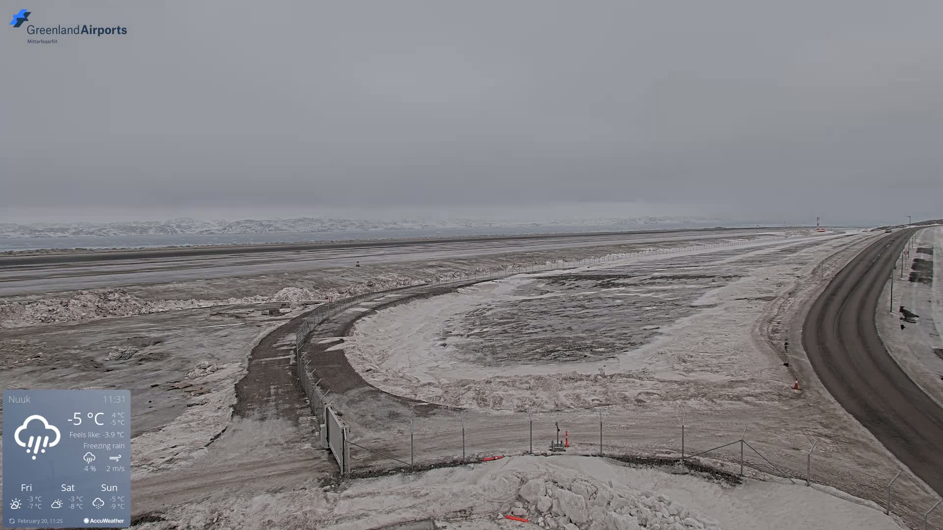 A snow-covered airport tarmac and road extend toward a distant, snow-dusted mountain range under an overcast, gray sky with freezing rain.