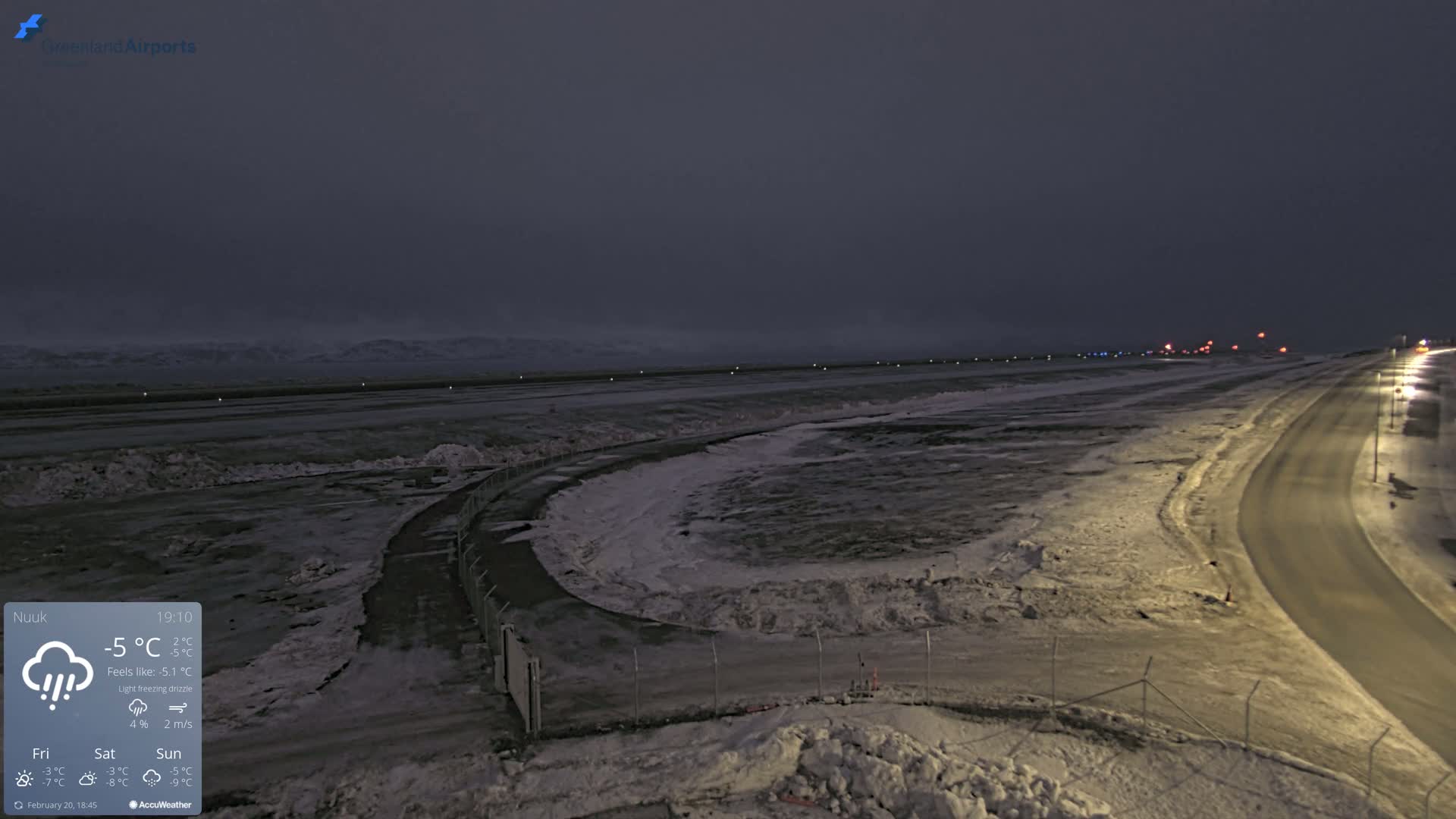 A snow-covered airport runway and road are visible under a dark, overcast sky with light freezing drizzle.