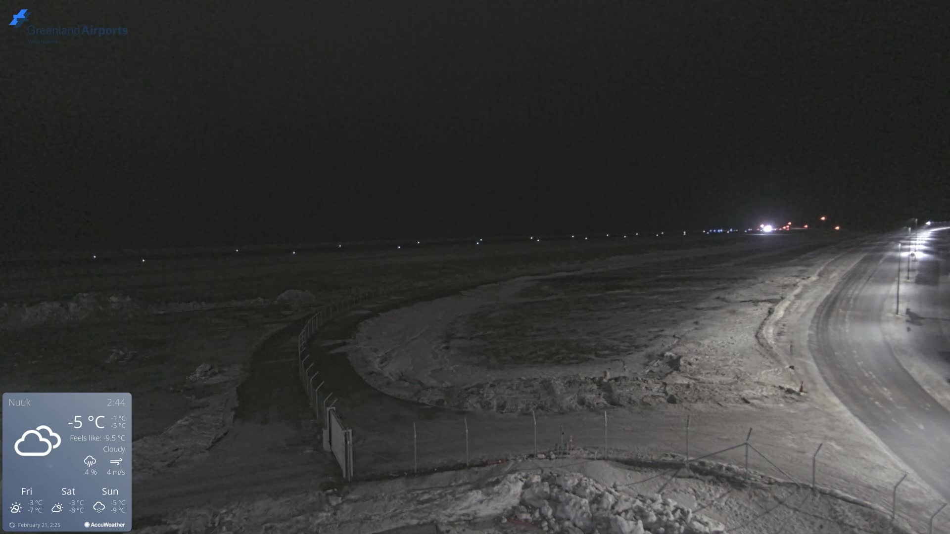 A nighttime, snowy, and cloudy scene shows a snow-covered road curving through a fenced-in area, with lights illuminating a distant airport runway.
