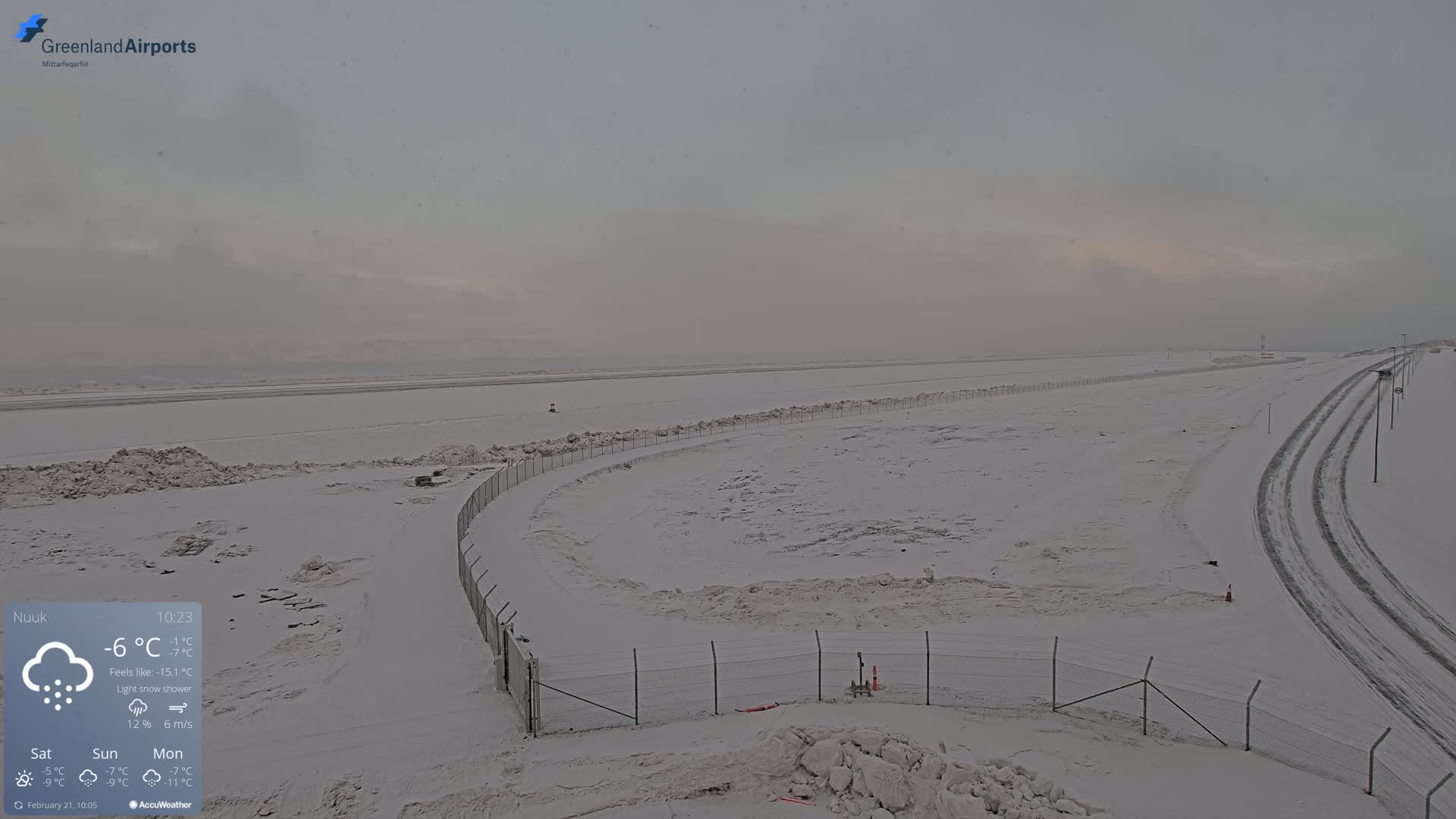 A snow-covered airport tarmac and surrounding area are visible under a light snow shower during overcast conditions.
