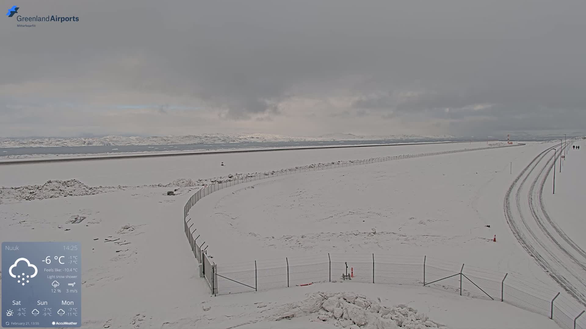A snow-covered airport runway and surrounding area with a light snow shower under an overcast sky.