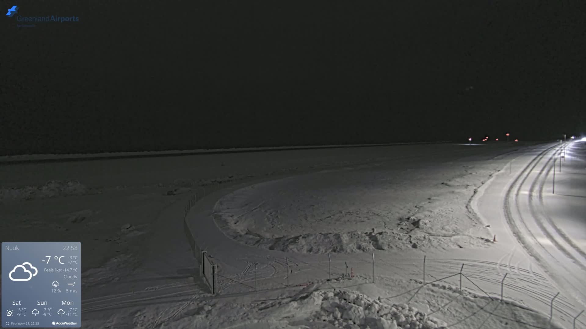 A snow-covered landscape at night under cloudy skies shows a curved road with tire tracks illuminated by streetlights, and a fenced-in area near a body of water.
