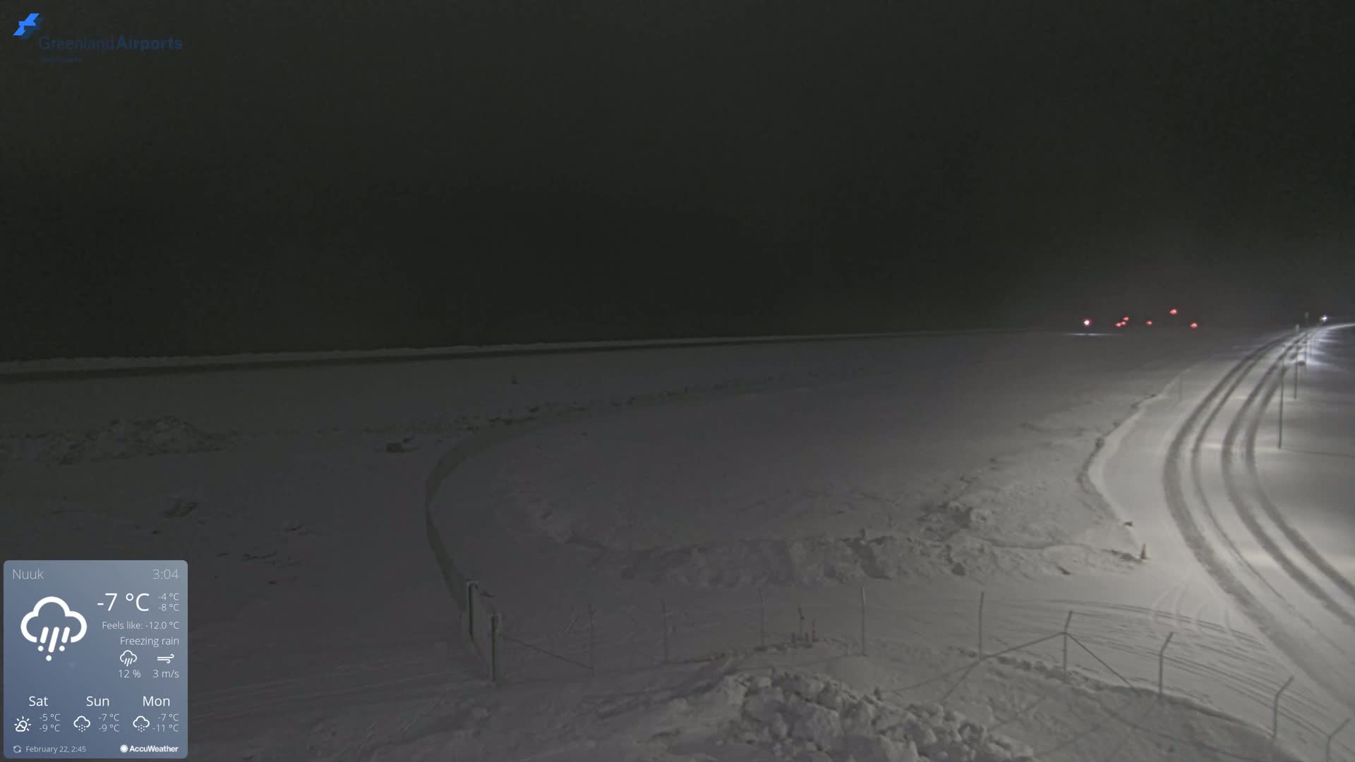 A snow-covered landscape at night under freezing rain conditions shows a road with tire tracks and faint vehicle lights in the distance.