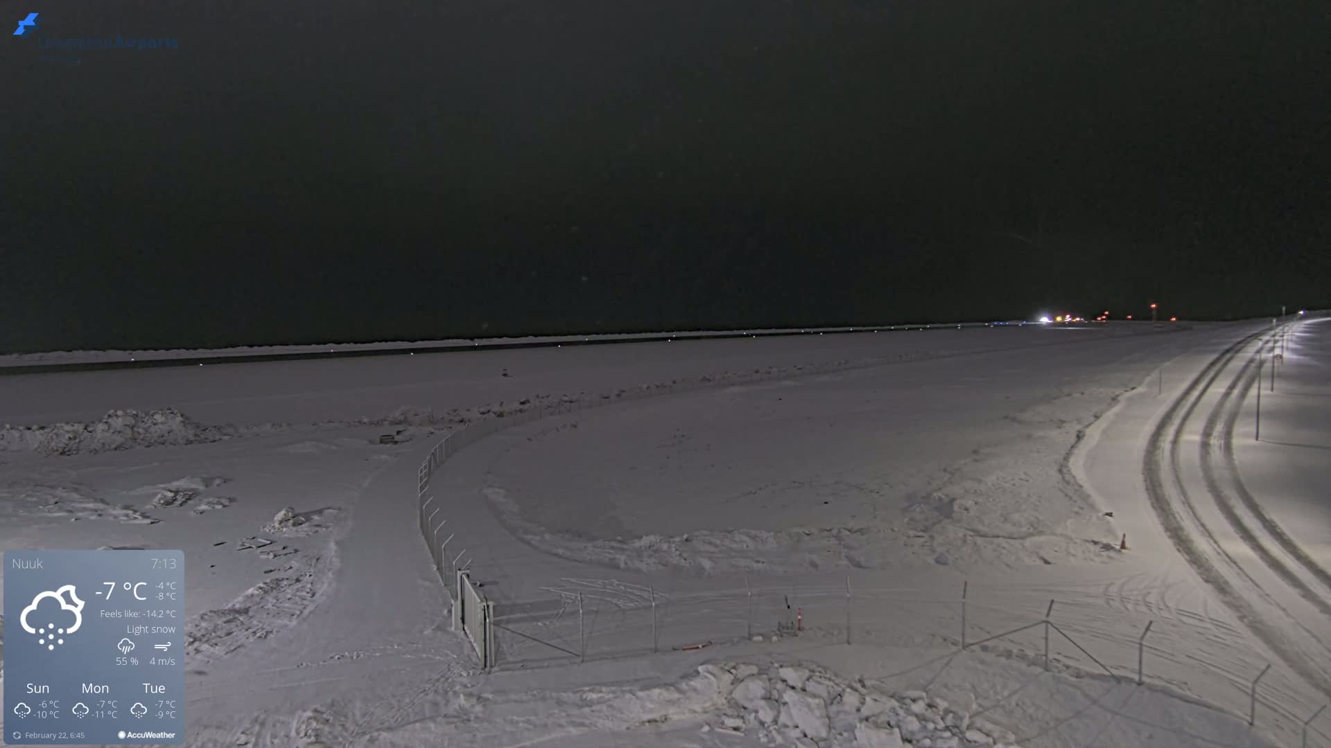 A snow-covered landscape at night under light snow, showing a road with tire tracks leading to a body of water partially visible in the distance.