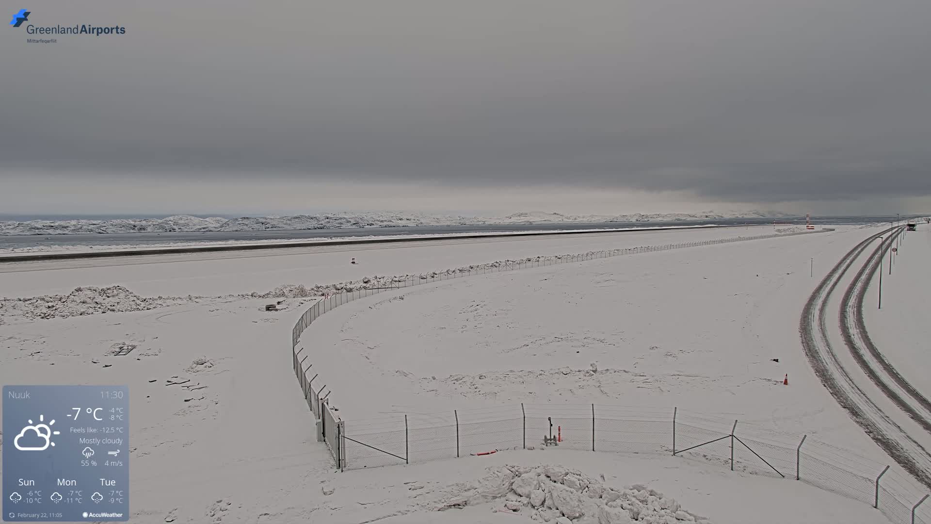 A snow-covered airport runway and surrounding area are visible under a mostly cloudy sky.