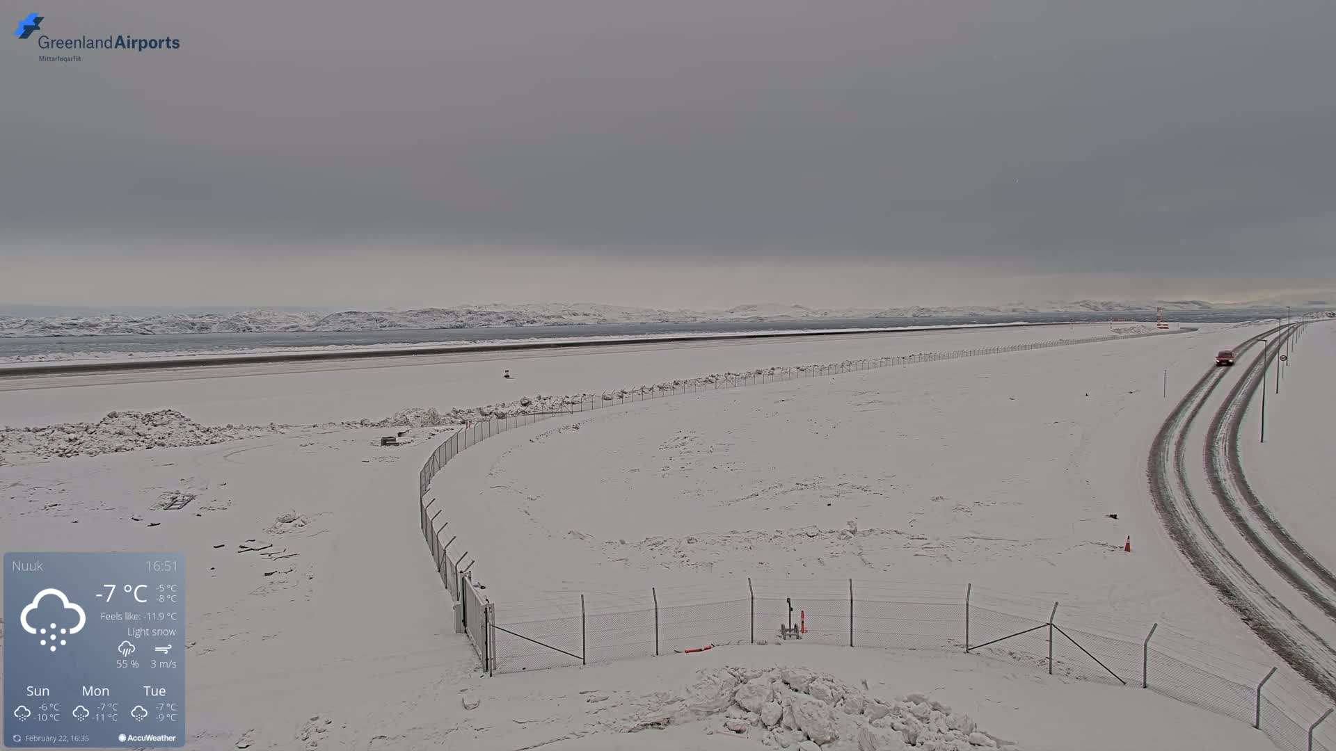 A snow-covered airport runway and surrounding area with a vehicle on a road in the distance under a gray, overcast sky; light snow is falling.