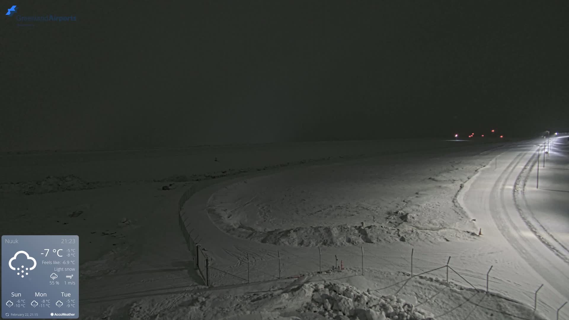 A snow-covered landscape at night with a road and fence visible under light snowfall.